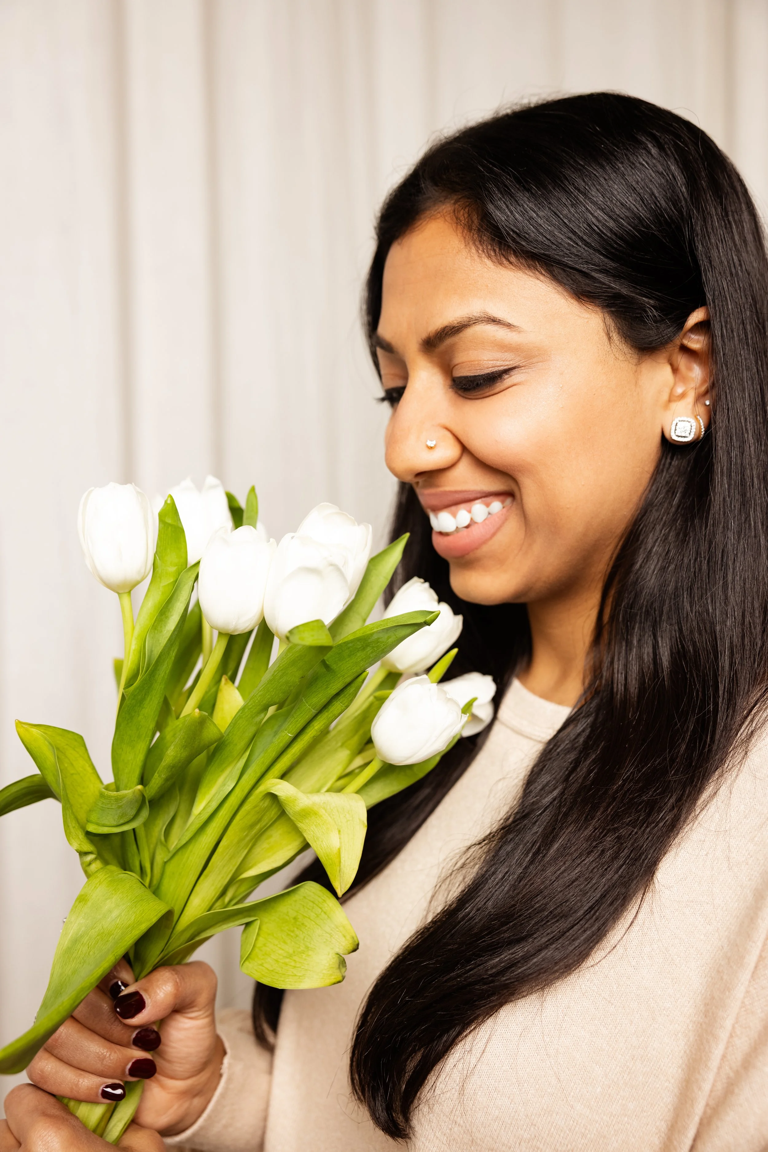 A woman with long dark hair, diamond earrings, and light skin is holding a bouquet of white tulips, smiling softly with her eyes closed, standing in front of a plain light-colored background.