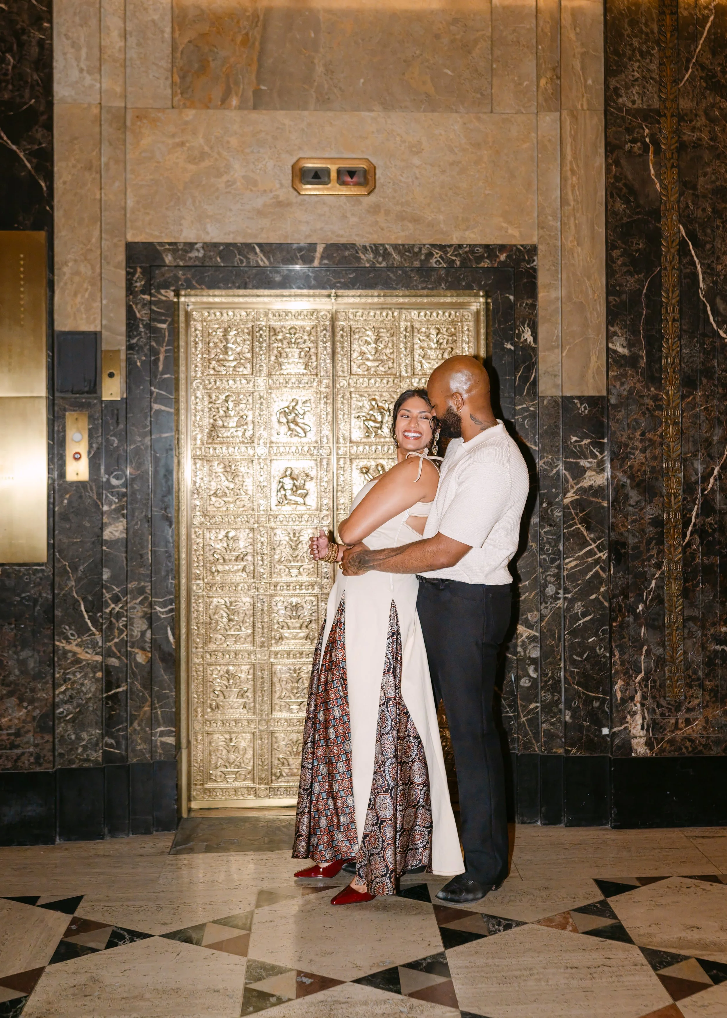 A couple standing in front of an ornate gold elevator door in a luxurious marble-floored lobby, smiling and embracing.