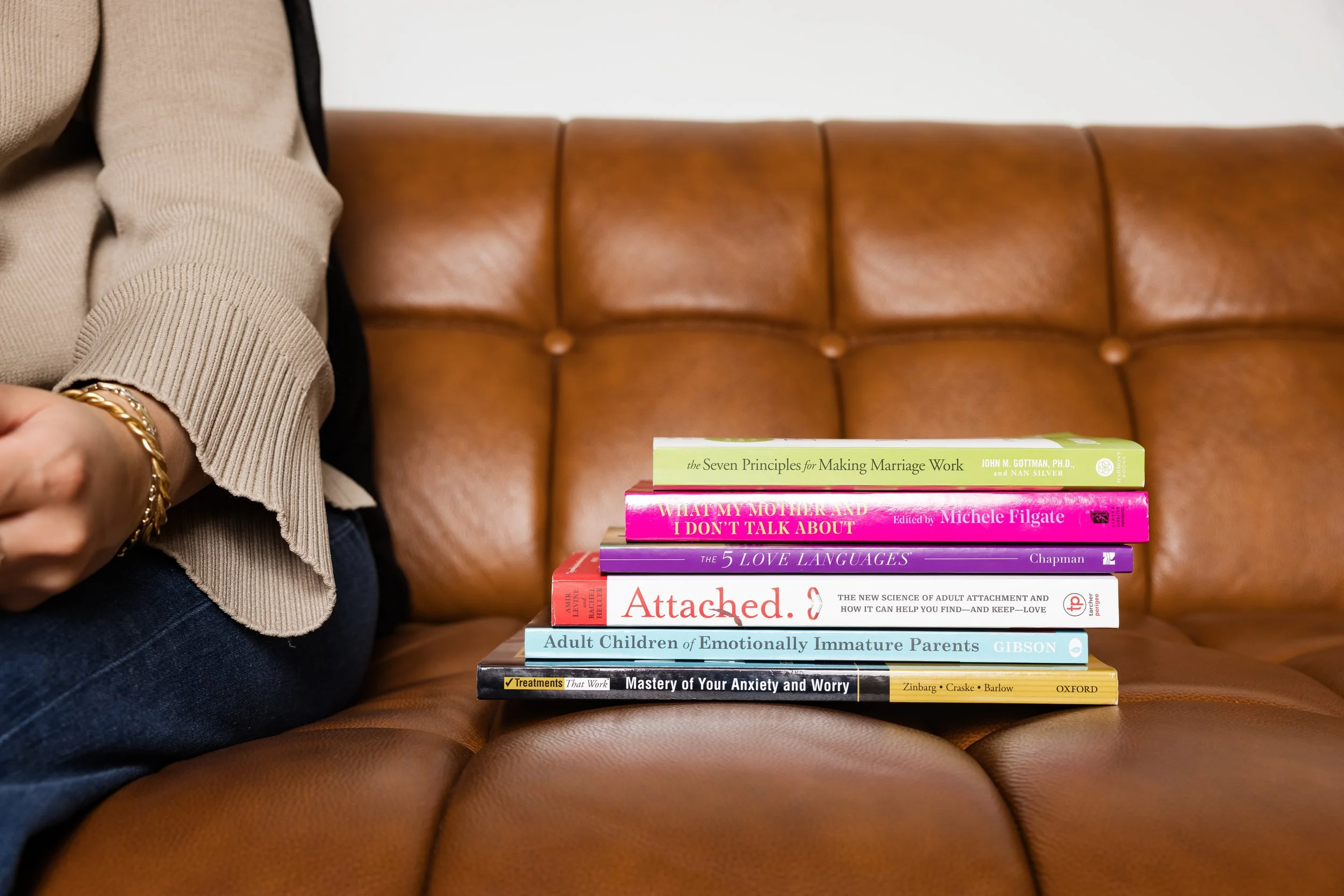 Part of a person's arm and torso on the left side, wearing a beige sweater and a gold bracelet, seated on a brown leather couch. In front, a stack of books on topics related to marriage, relationships, communication, and emotional health, with a plain white wall in the background.