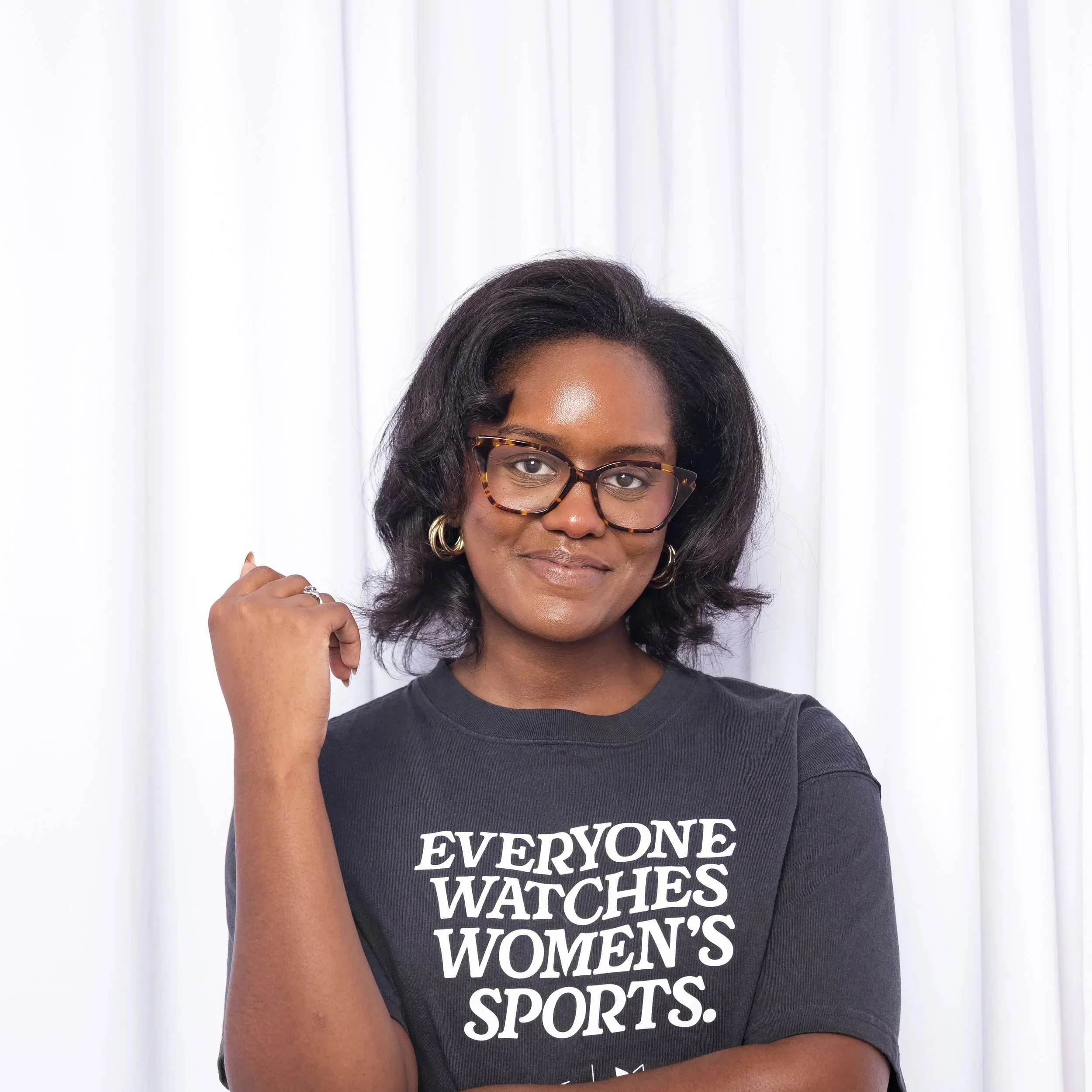 A woman with dark hair and glasses, wearing a black shirt with white text that says 'EVERYONE WATCHES WOMEN'S SPORTS,' stands in front of white curtains.