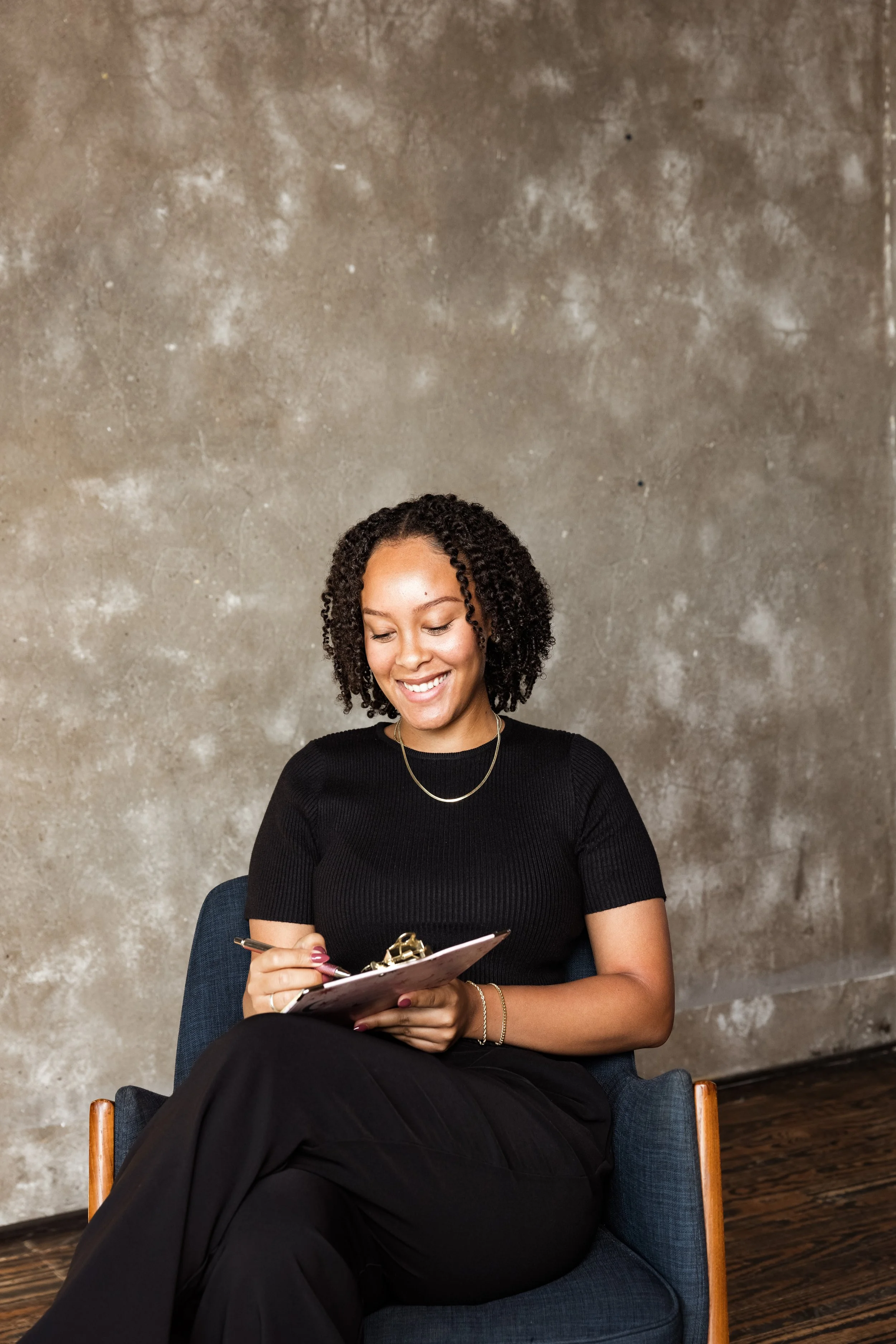 Ayesha Youngblood smiling and writing on a clipboard, seated in a blue armchair in front of a textured gray wall.