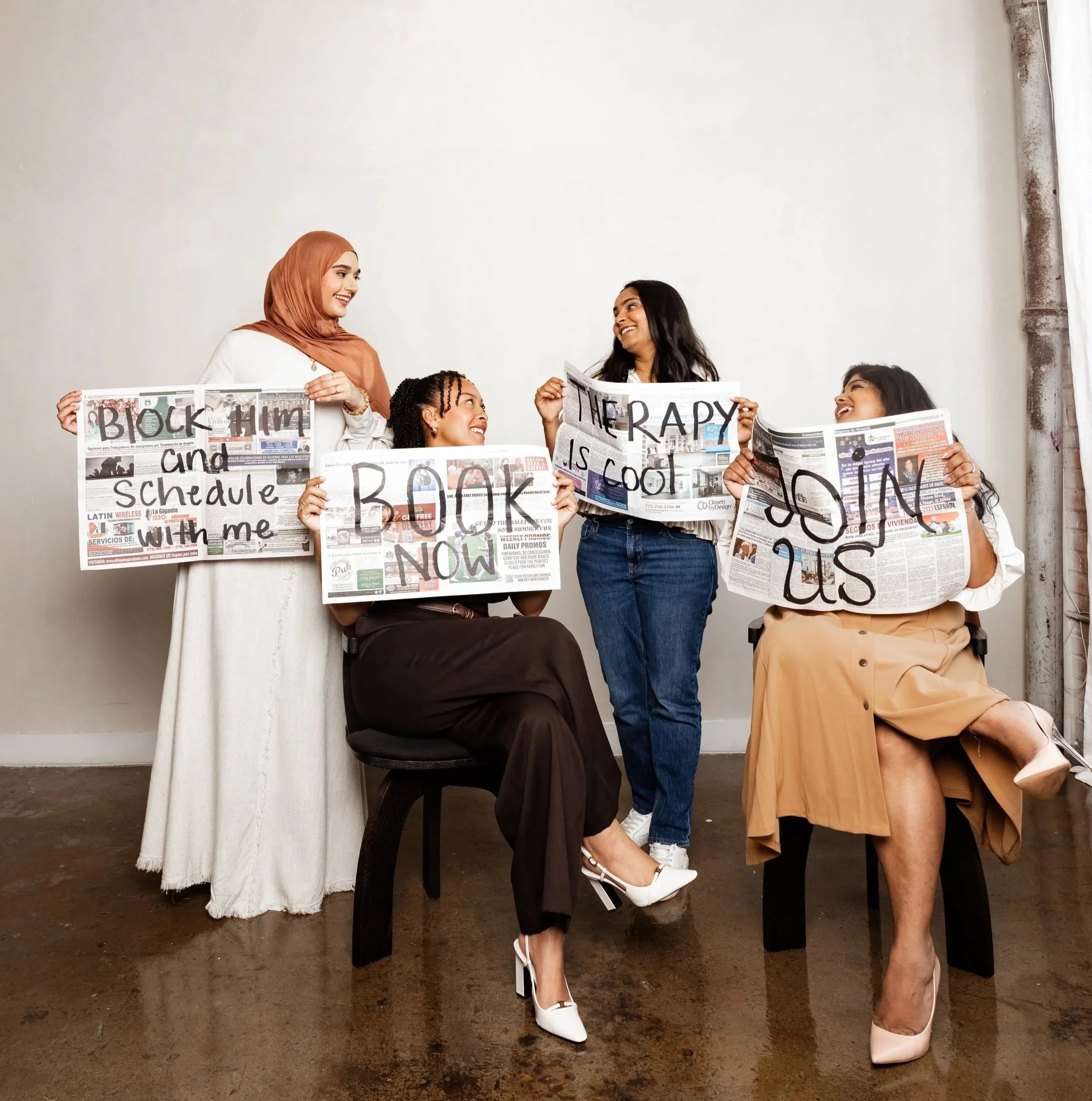 Group of women holding newspapers while sitting and standing in front of a plain wall. They are smiling and interacting with each other.