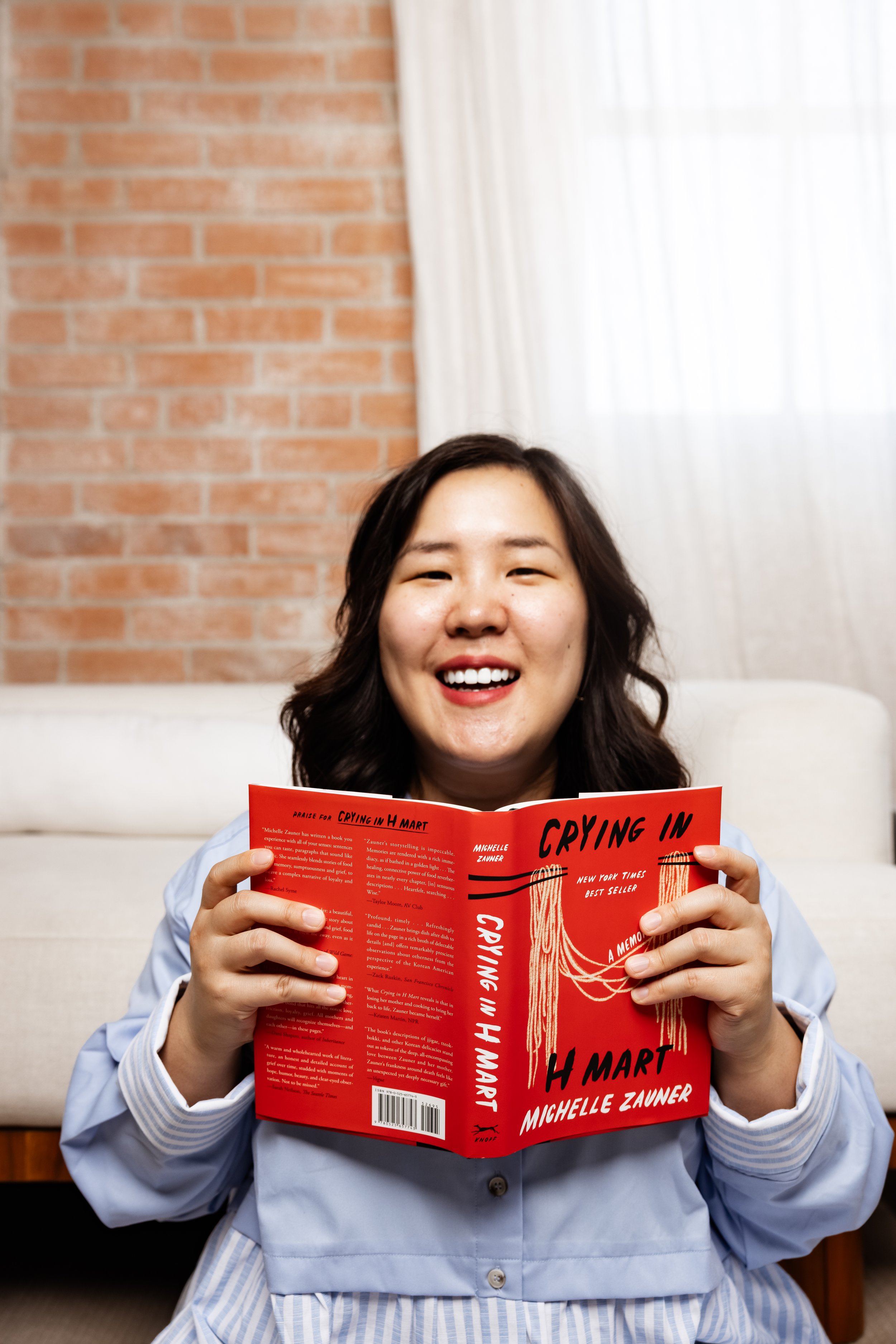 A woman with long dark hair wearing a light blue and white striped shirt smiling and holding a red book titled 'Crying in H Mart' by Michelle Zauner, in a room with a brick wall and a window with white curtains.