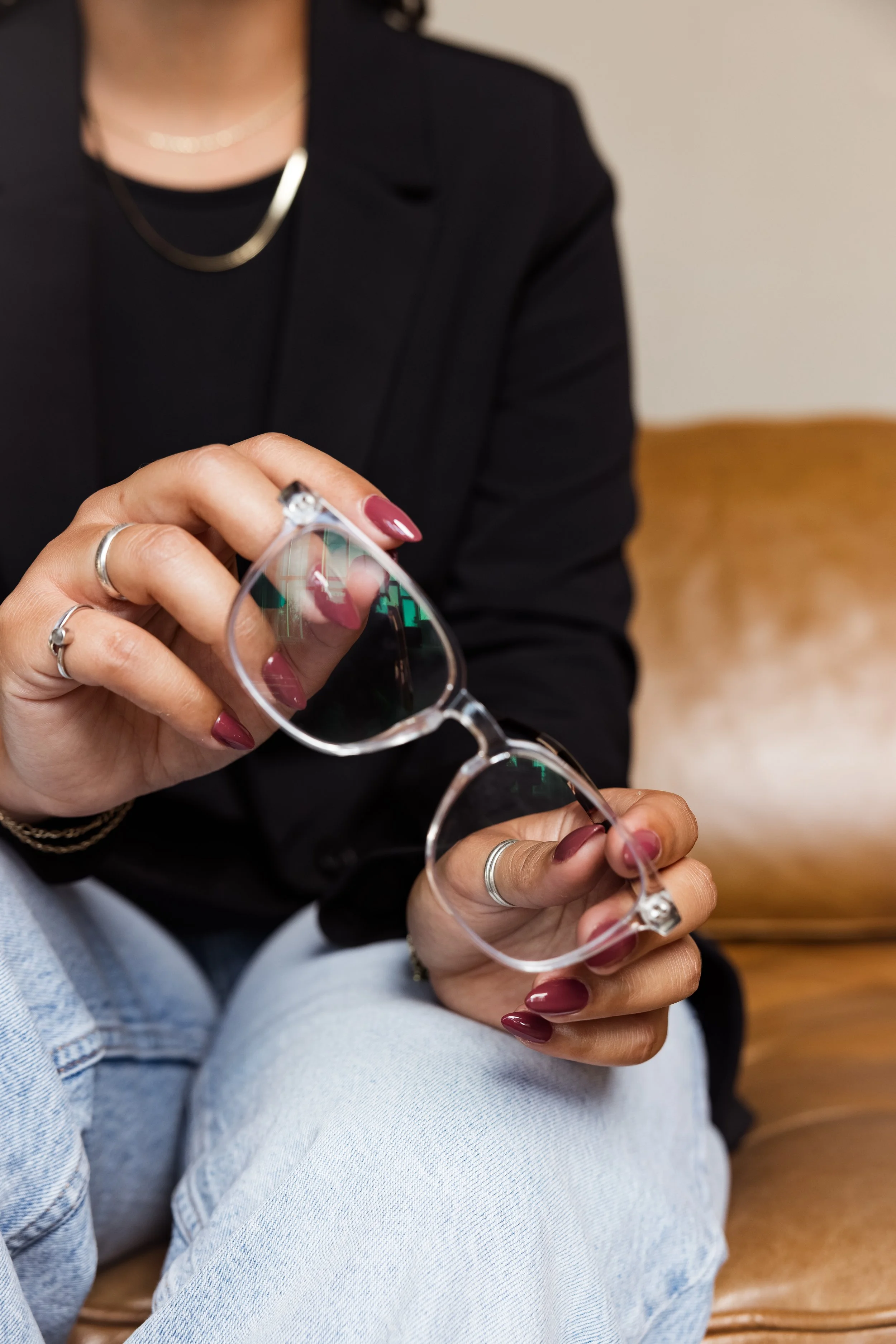 Close-up of a person wearing a black jacket and jewelry, sitting on a wooden chair, holding transparent glasses with a reflective surface and manicure with maroon nails.