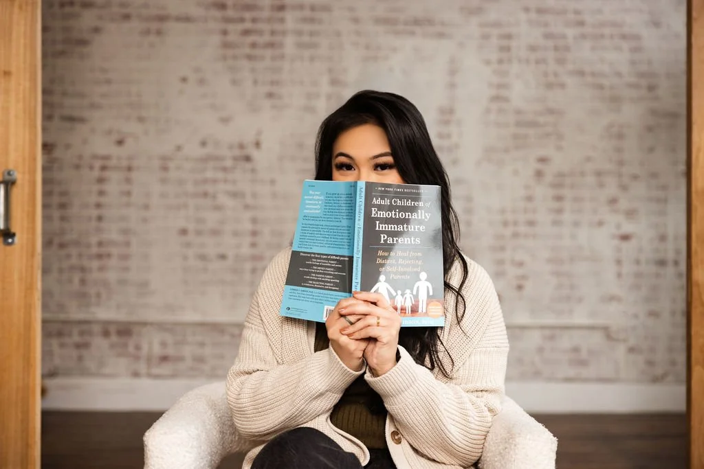 A woman sitting on a cozy chair, holding a book titled "Adult Children of Emotionally Immature Parents" in front of her face, with her eyes peeking over the top of the book.