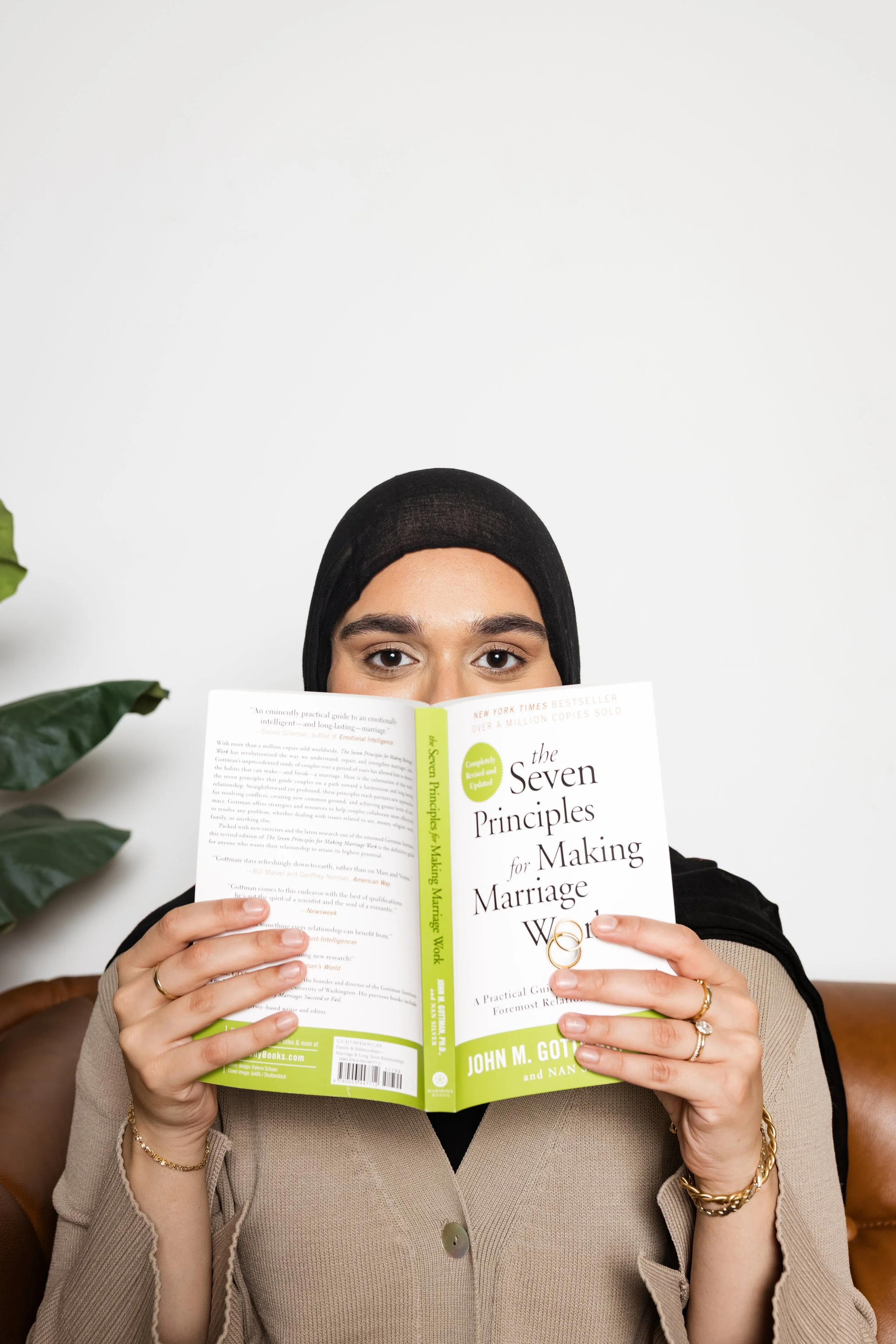 A woman with a beige blazer and gold jewelry is sitting and holding a book titled 'The Seven Principles for Making Marriage Work' by John M. Gottman, partially covering her face, with only her eyes and forehead visible.