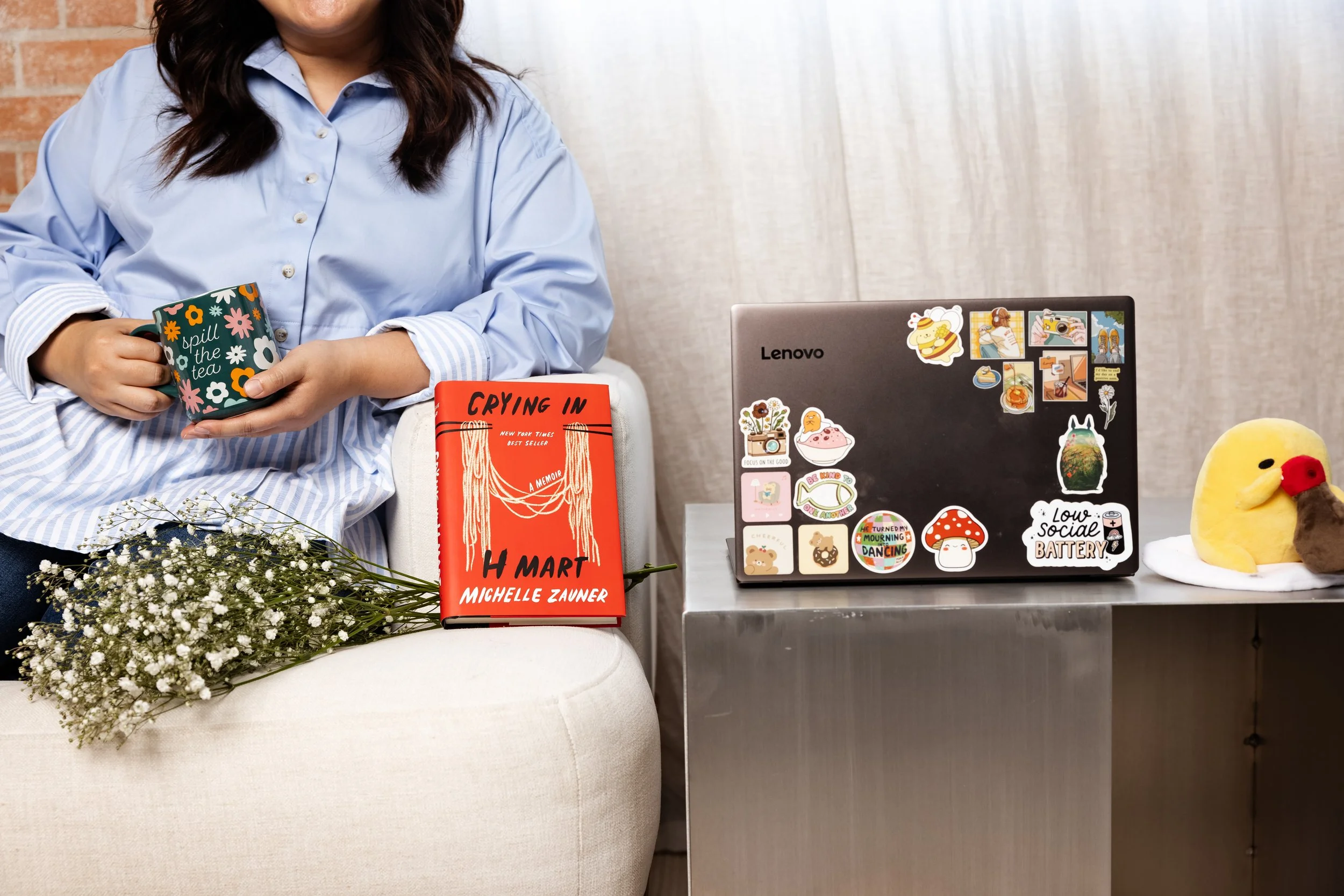 A woman sitting on a white chair holding a floral mug that reads 'spill the tea', with a bouquet of baby's breath flowers on her lap, a red book titled 'Crying in H Mart' by Michelle Zauner resting on her knee, a silver table with a decorated laptop,