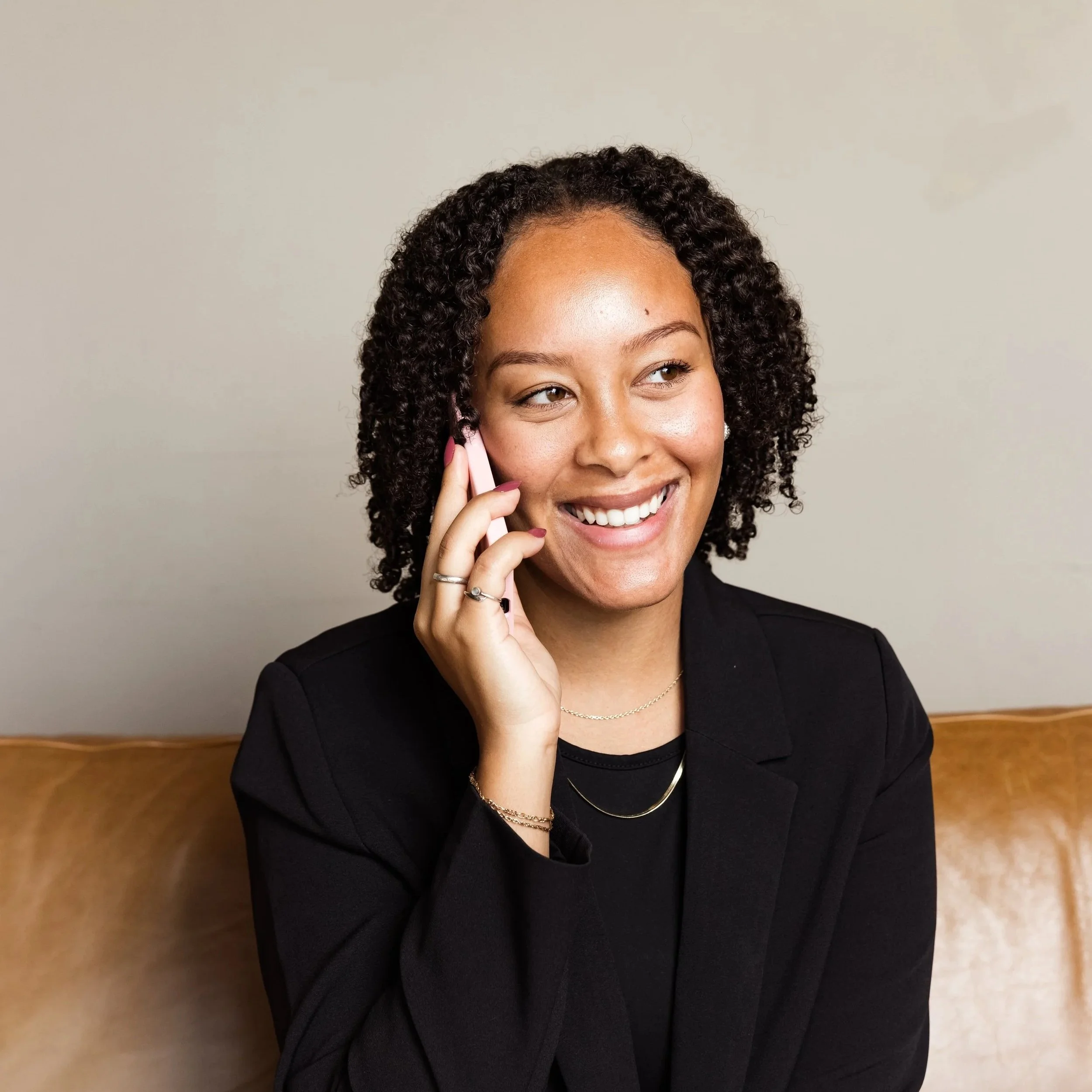 A woman with curly dark hair and a warm smile, talking on a pink phone, wearing a black blazer, layered necklaces, and rings, standing in front of a light grey wall with a wooden baseboard.