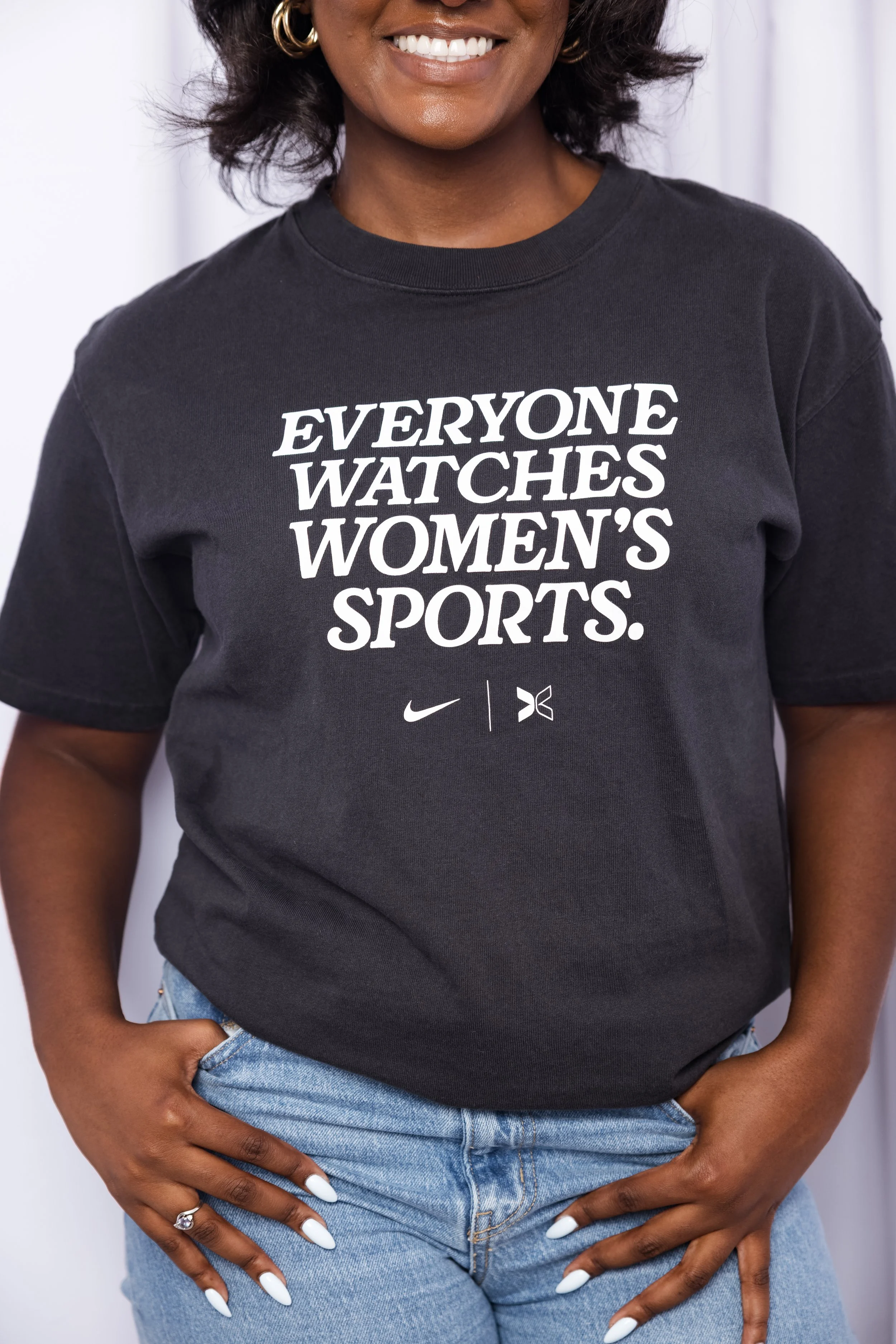 Close-up of a person wearing a black T-shirt with the message "Everyone Watches Women's Sports" and logos for Nike and another brand. The person has dark skin, a gold hoop earring, and long, pointed white nails. They are standing with their hands on their hips, wearing light blue jeans.