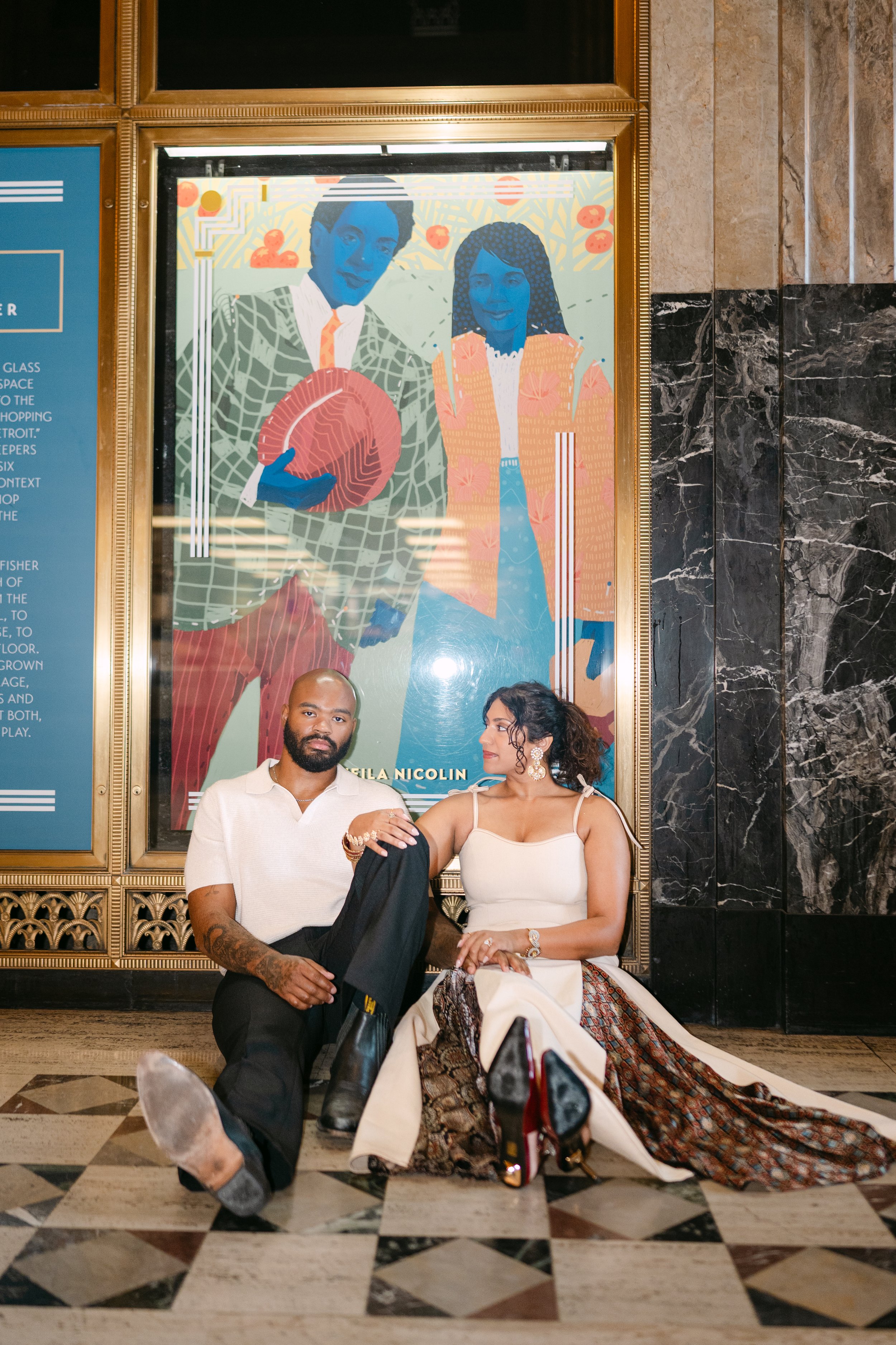 Two people sitting on the floor in front of a large colorful poster. The man has a beard, tattoos, and is wearing a white shirt and black pants, holding a black jacket and a plastic bag. The woman is wearing a white top and a patterned skirt, holding a handbag. They are inside a building with marble walls and a checkered floor.