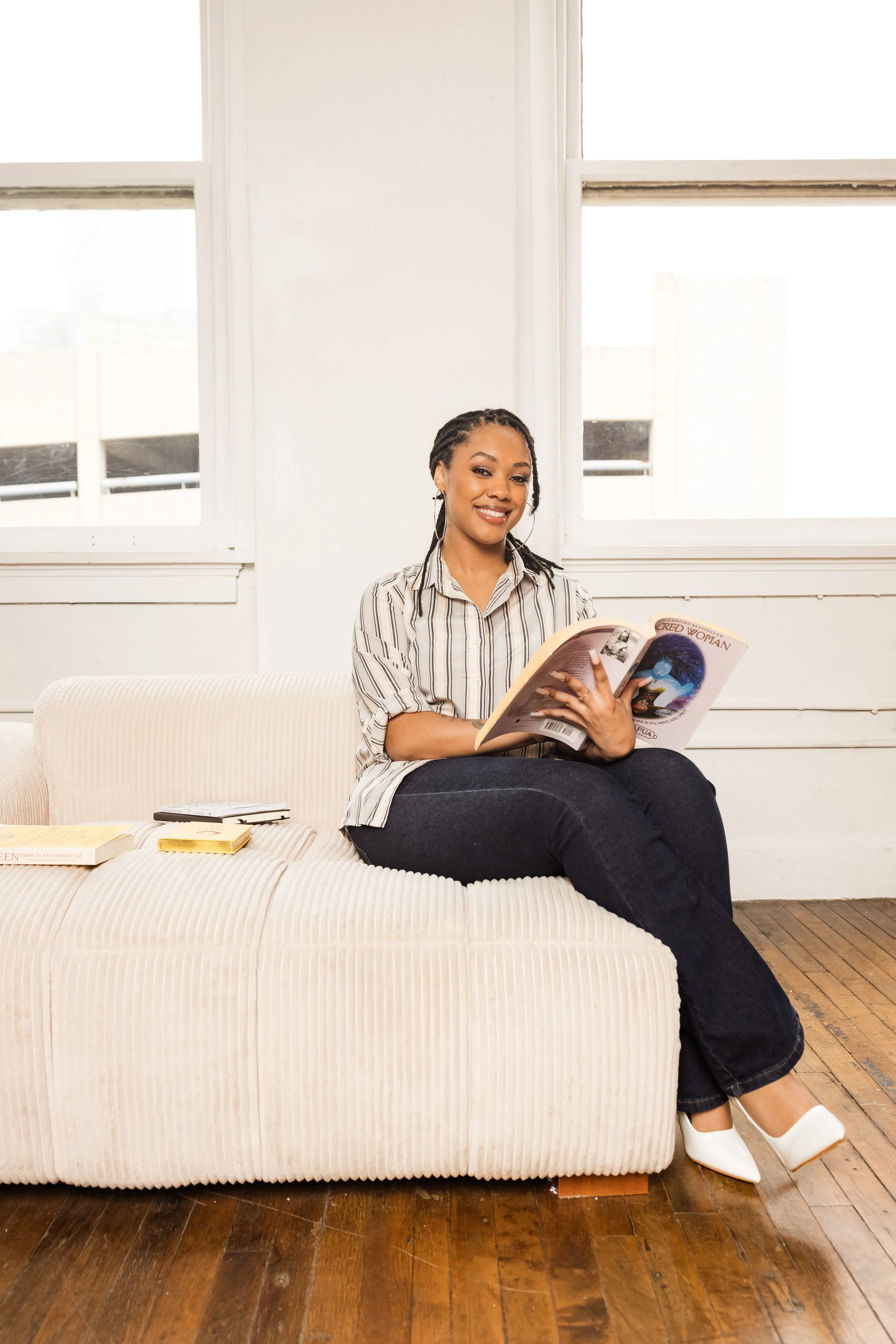 A woman sitting on a beige upholstered sofa, smiling and holding a magazine titled 'Woman'. She is wearing a striped shirt, dark jeans, and white shoes. There are books on the sofa and natural light coming through front windows.