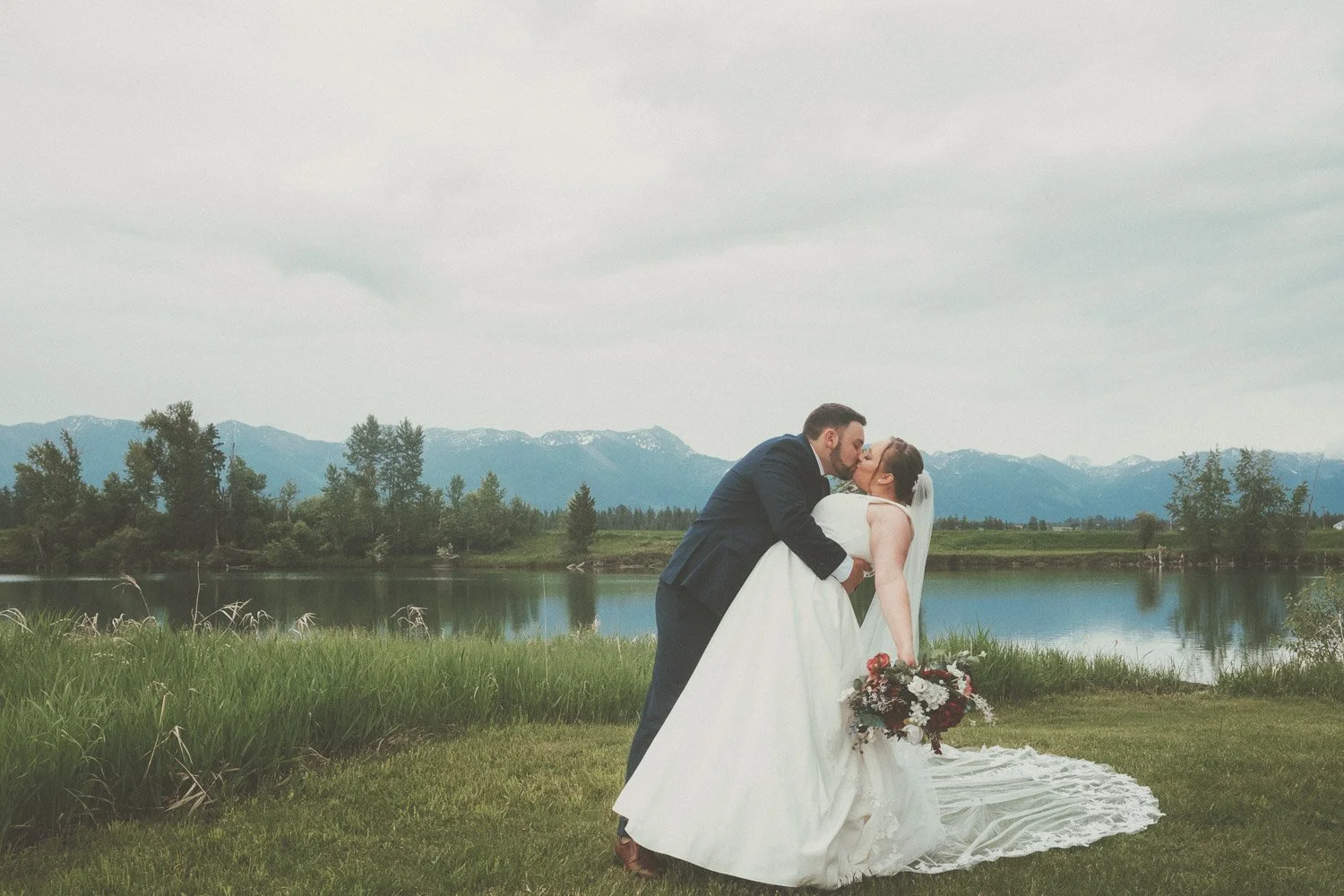 A newlywed couple sharing a kiss outdoors near a lake with mountains in the background. The groom is dressed in a dark suit and the bride in a white wedding gown holding a bouquet of flowers.