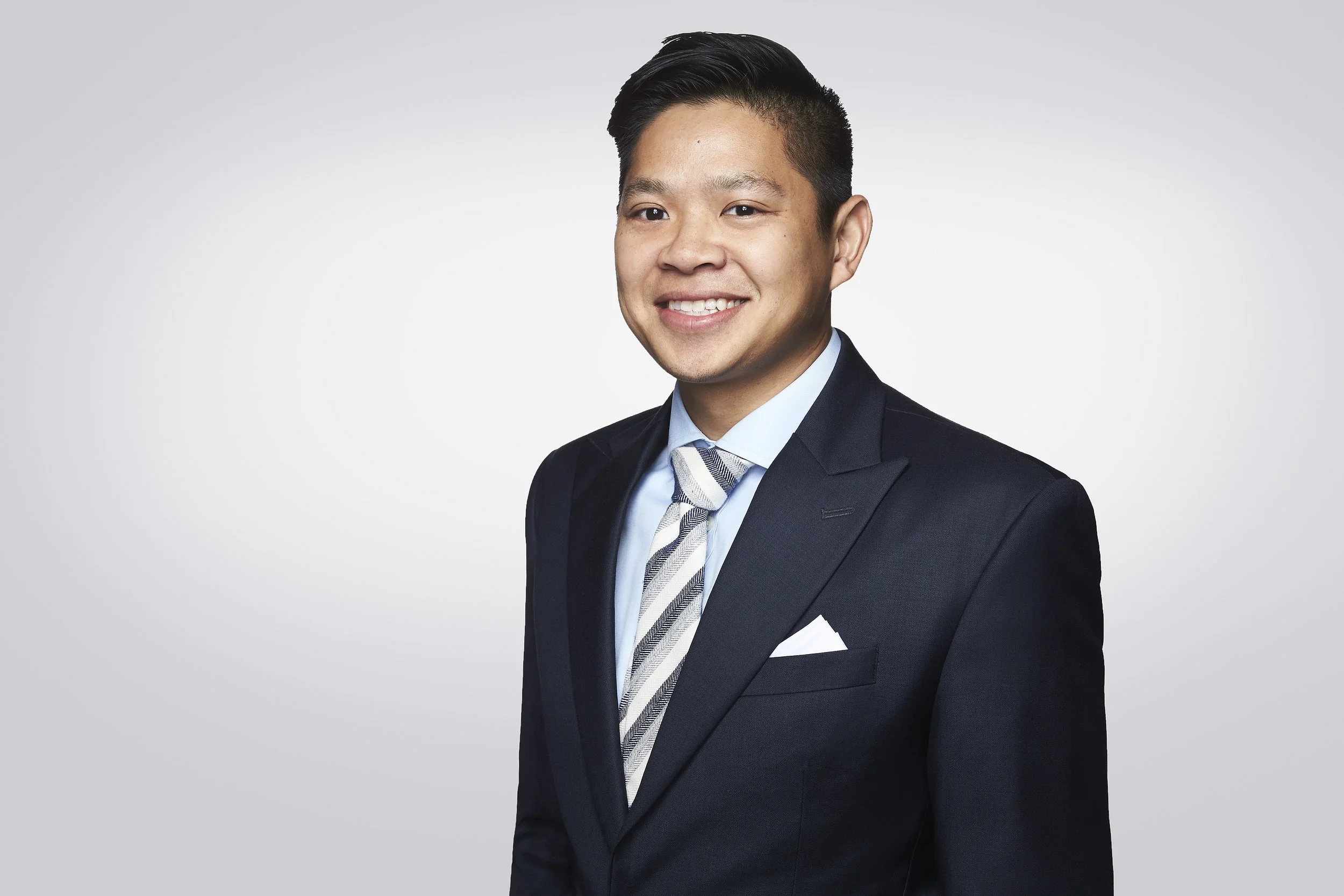 A young Asian man in a dark suit, light blue dress shirt, striped tie, with a white pocket square, smiling at the camera against a gray gradient background.