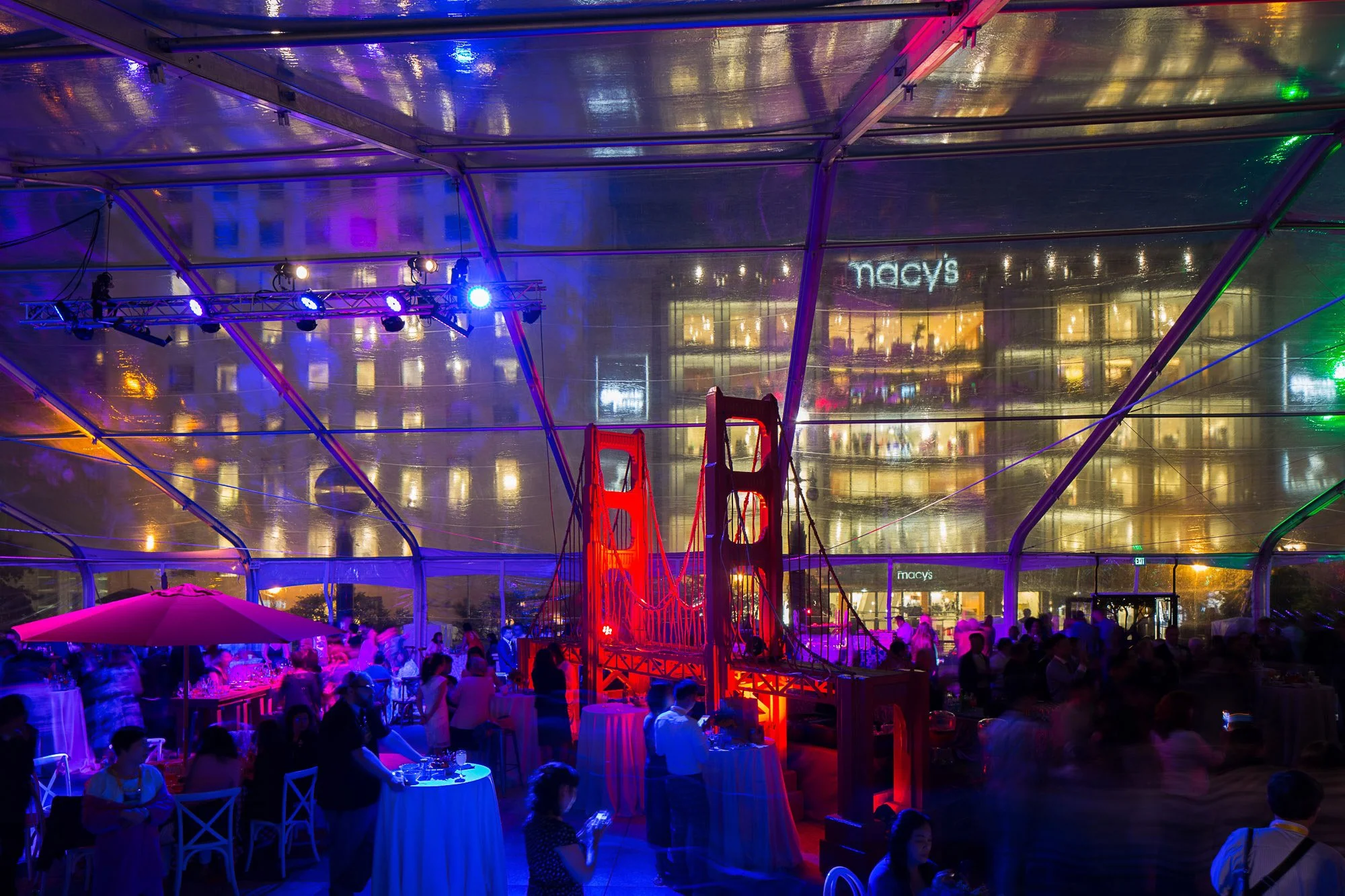 Nighttime scene at an indoor event with colorful lighting, a red suspension bridge replica, tables with people dining, and the Macy's store visible through the glass ceiling.