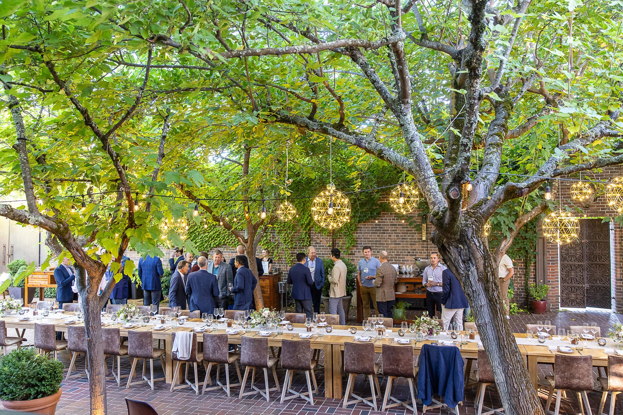 A lively outdoor gathering under trees with hanging spherical light fixtures. People are socializing near a long dining table set with plates, glasses, and floral centerpieces, against a brick wall decorated with green vines.