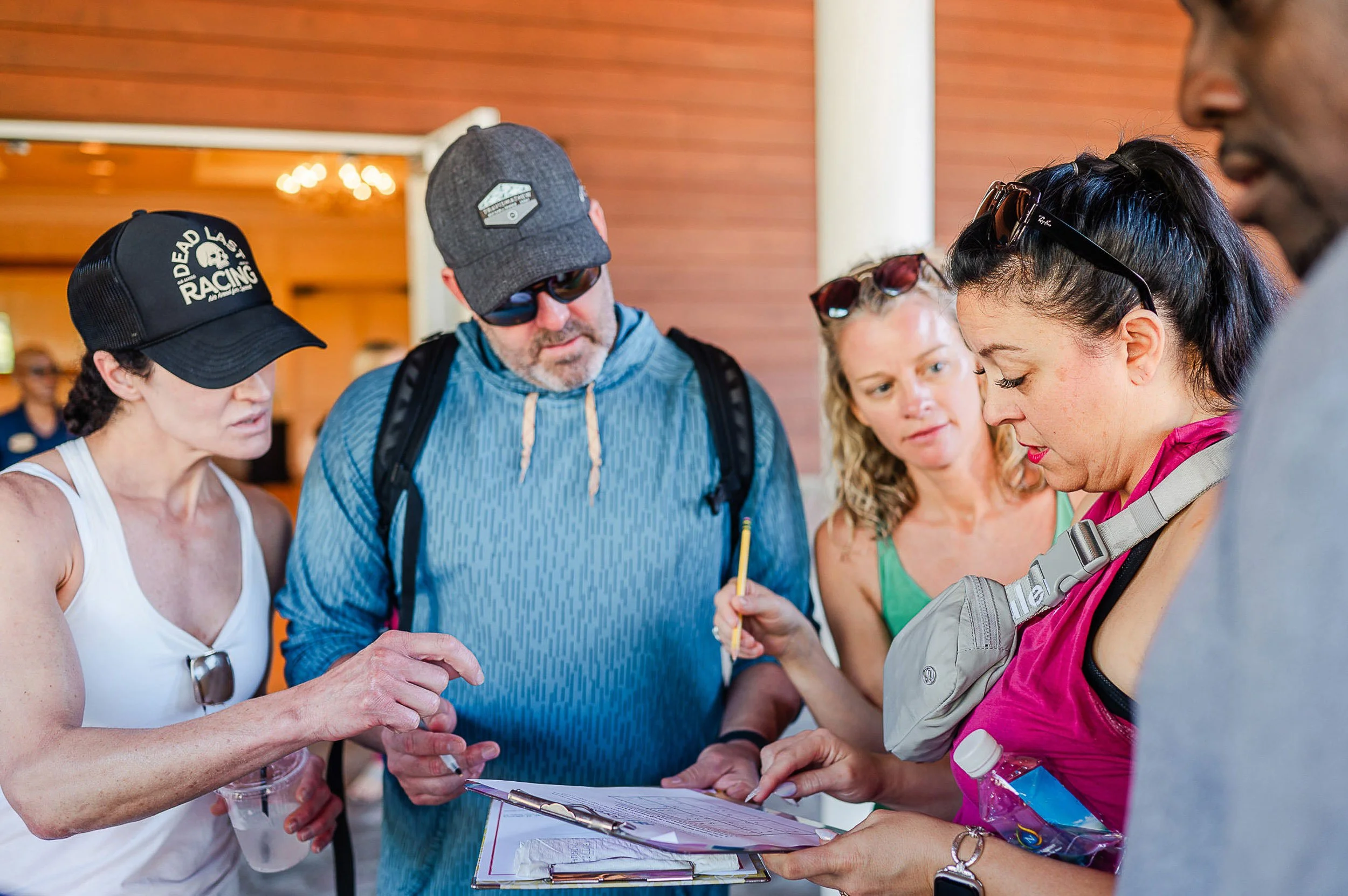 A group of people gathered around looking at a clipboard with papers, outdoors with a wood-paneled background.