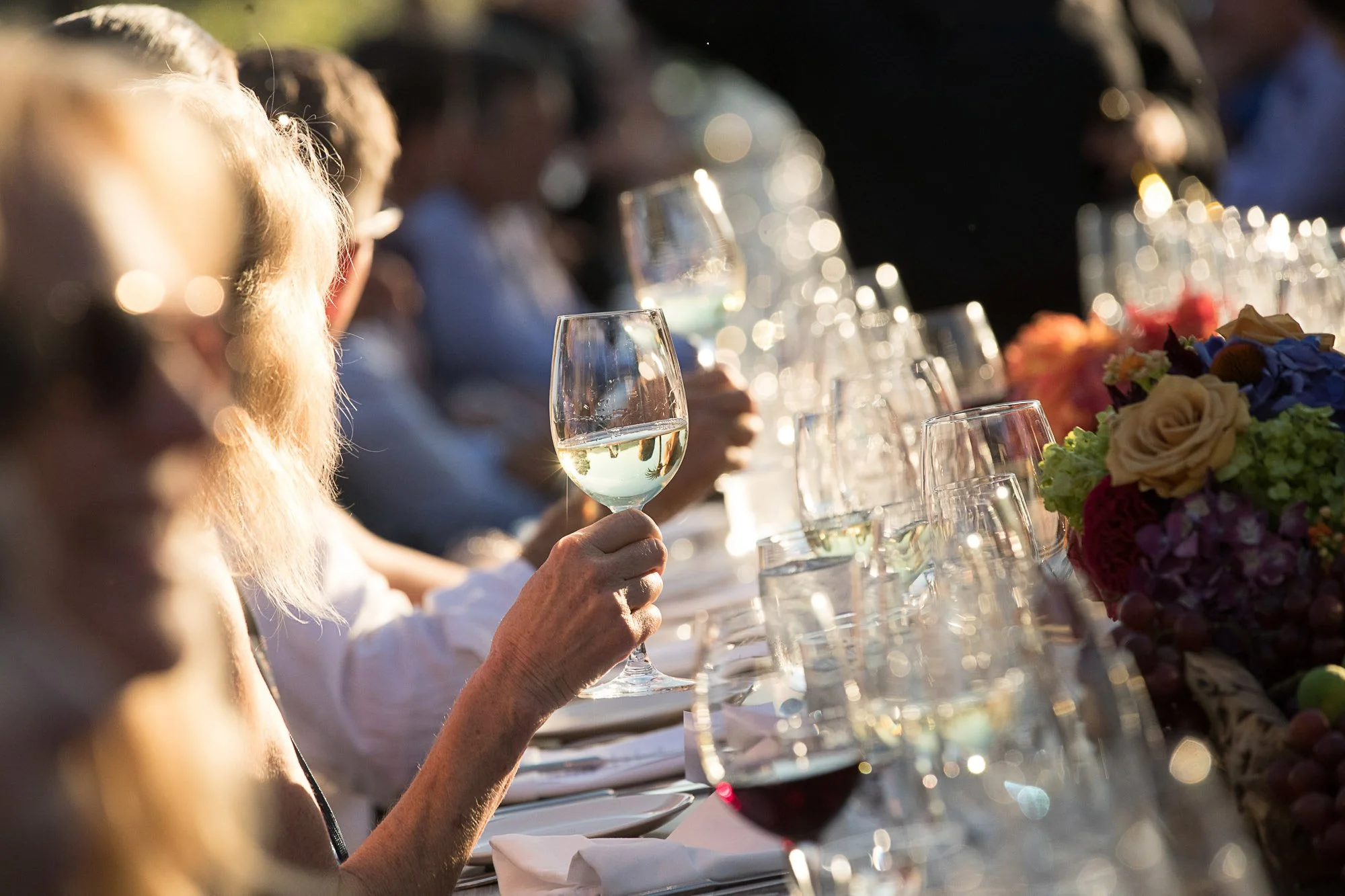 People sitting at a long table outdoors, celebrating with wine glasses and floral arrangements, illuminated by warm sunlight.