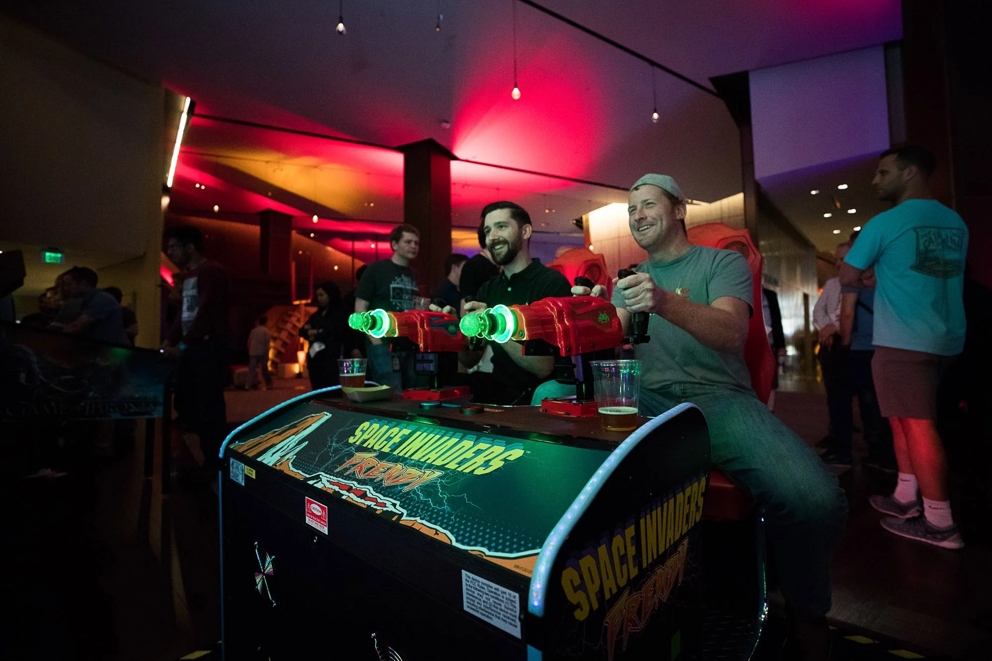 Two smiling men playing a space-themed pinball game at an arcade, with colorful neon lights overhead and other people in the background.