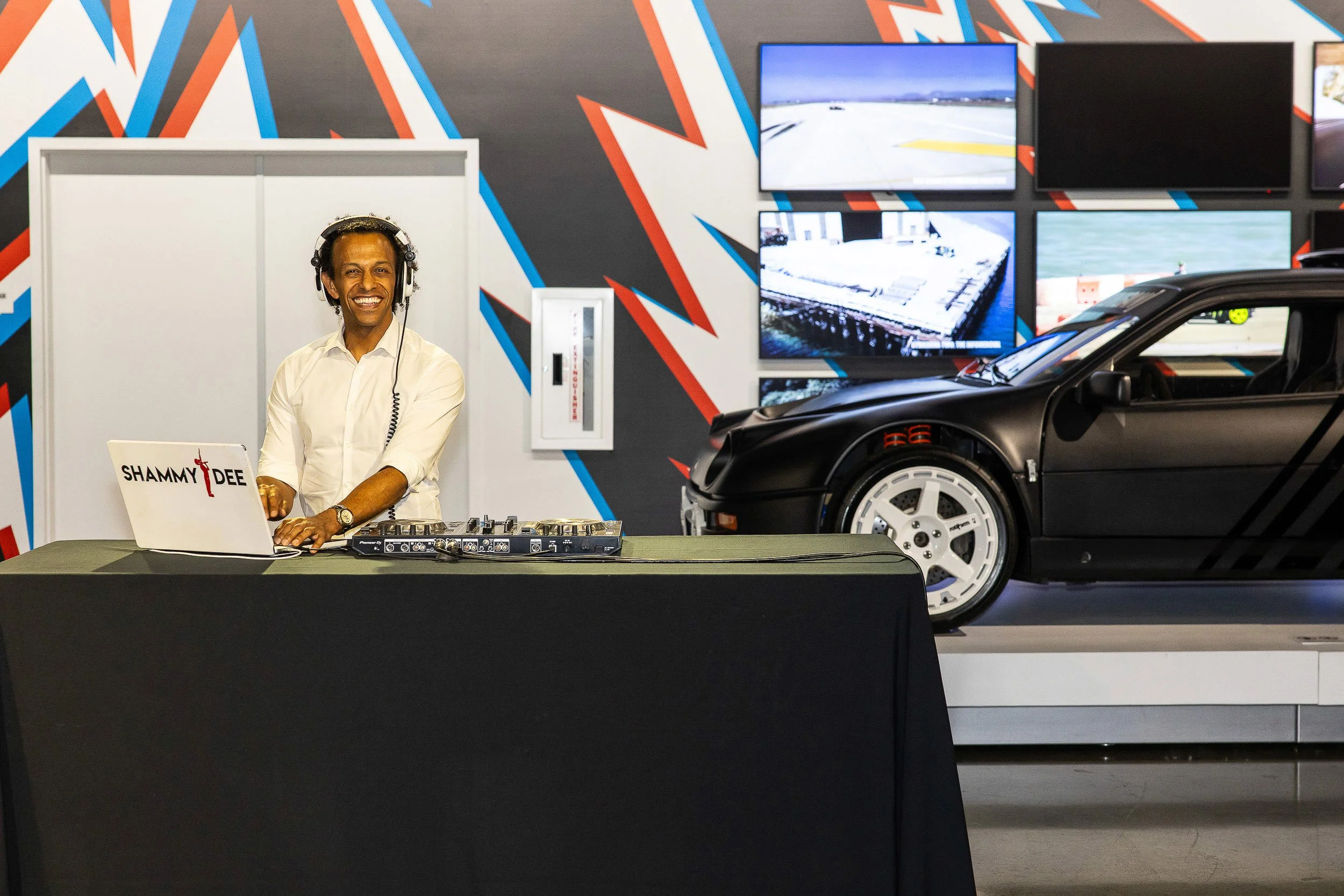 A man wearing headphones and a white shirt smiling at a DJ setup in front of a wall with colorful geometric designs. Behind him are multiple television screens displaying various images, including a race track and a sports stadium. To the right, part