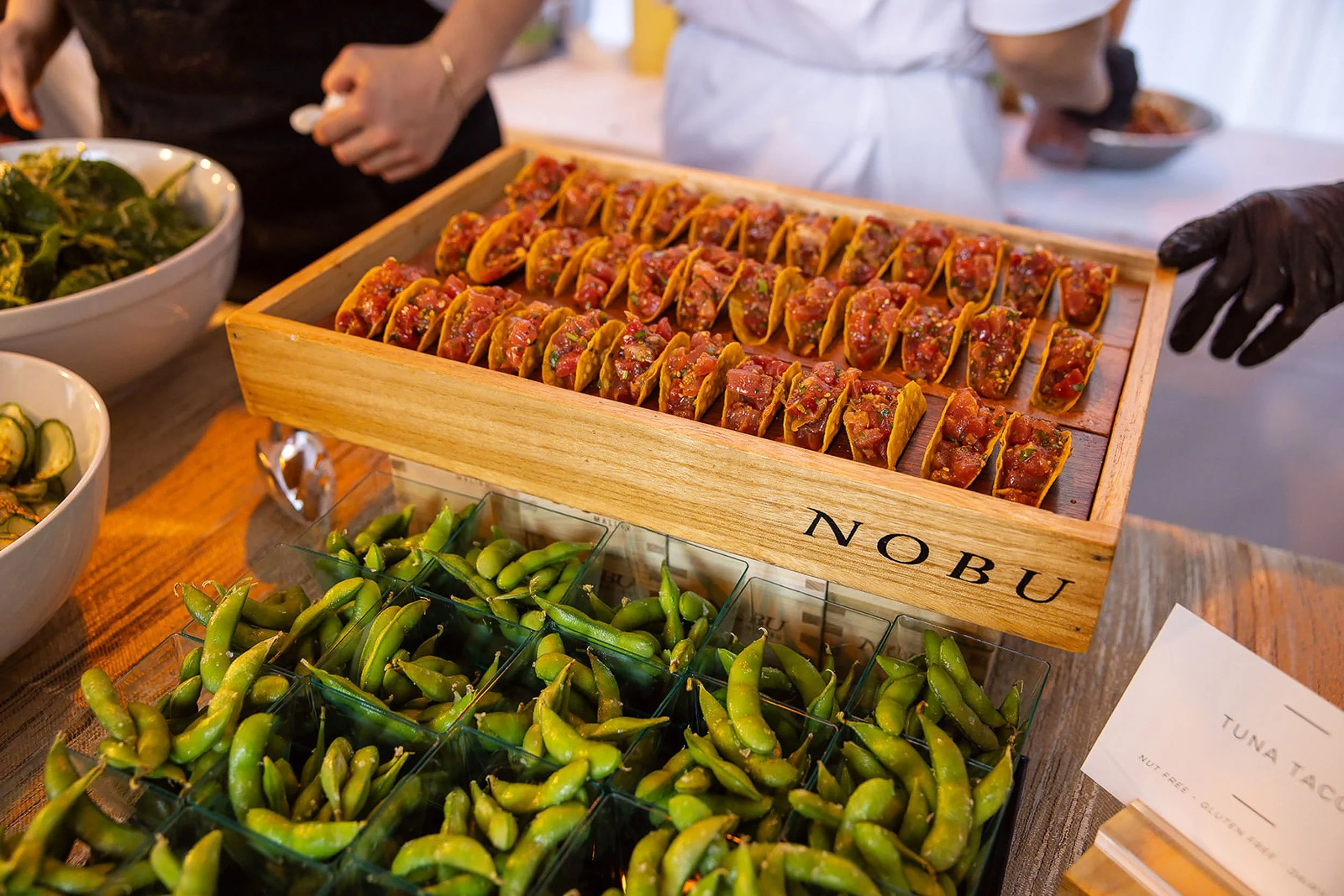 A tray of tacos with diced meat and toppings on a wooden stand, surrounded by bowls of salad and edamame at a food event.