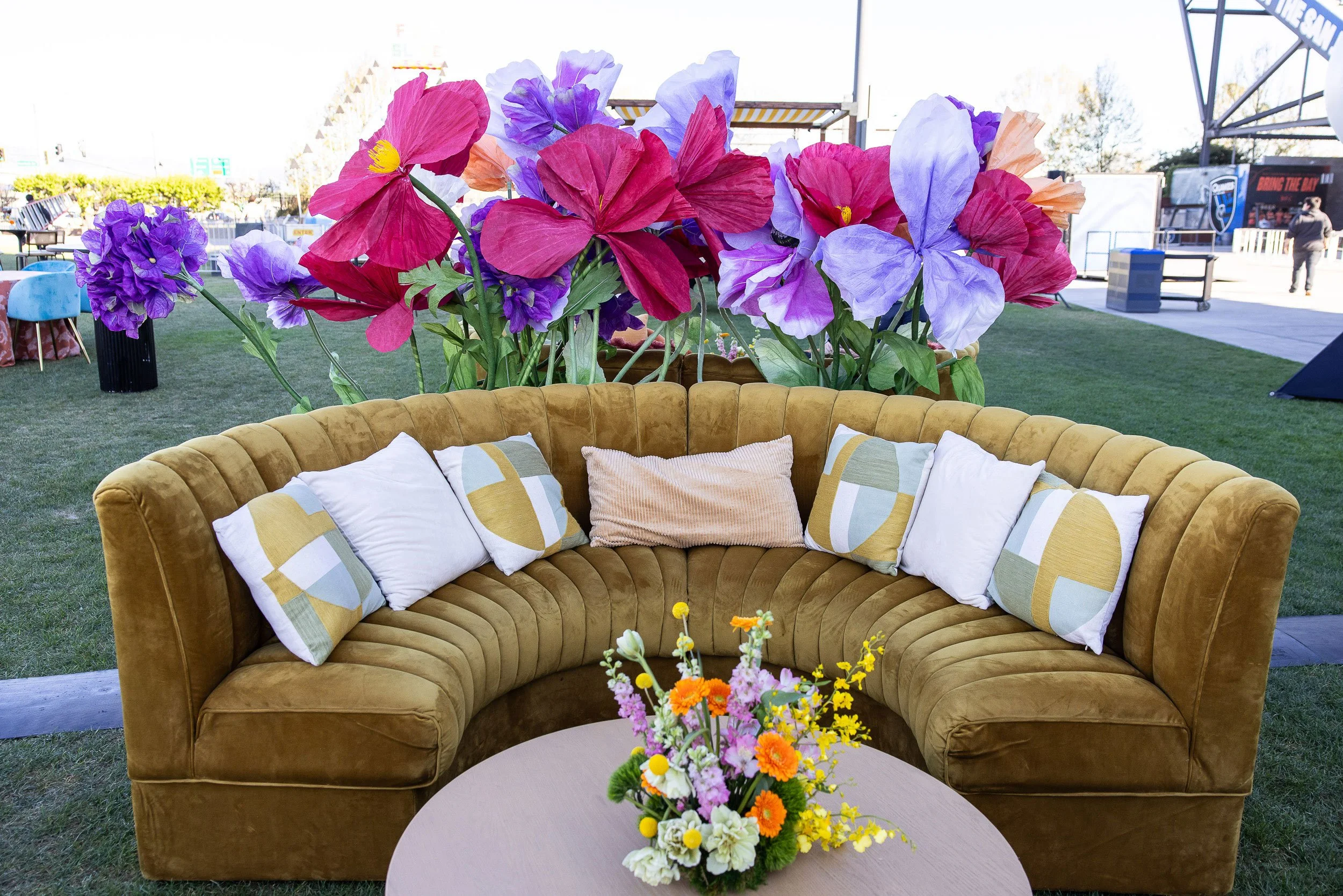A mustard-colored velvet mohair curved sofa with several patterned pillows, placed outdoors on a grassy area during daytime. Behind the sofa, there is a large floral arrangement made of colorful artificial flowers, including pink, purple, and lavende