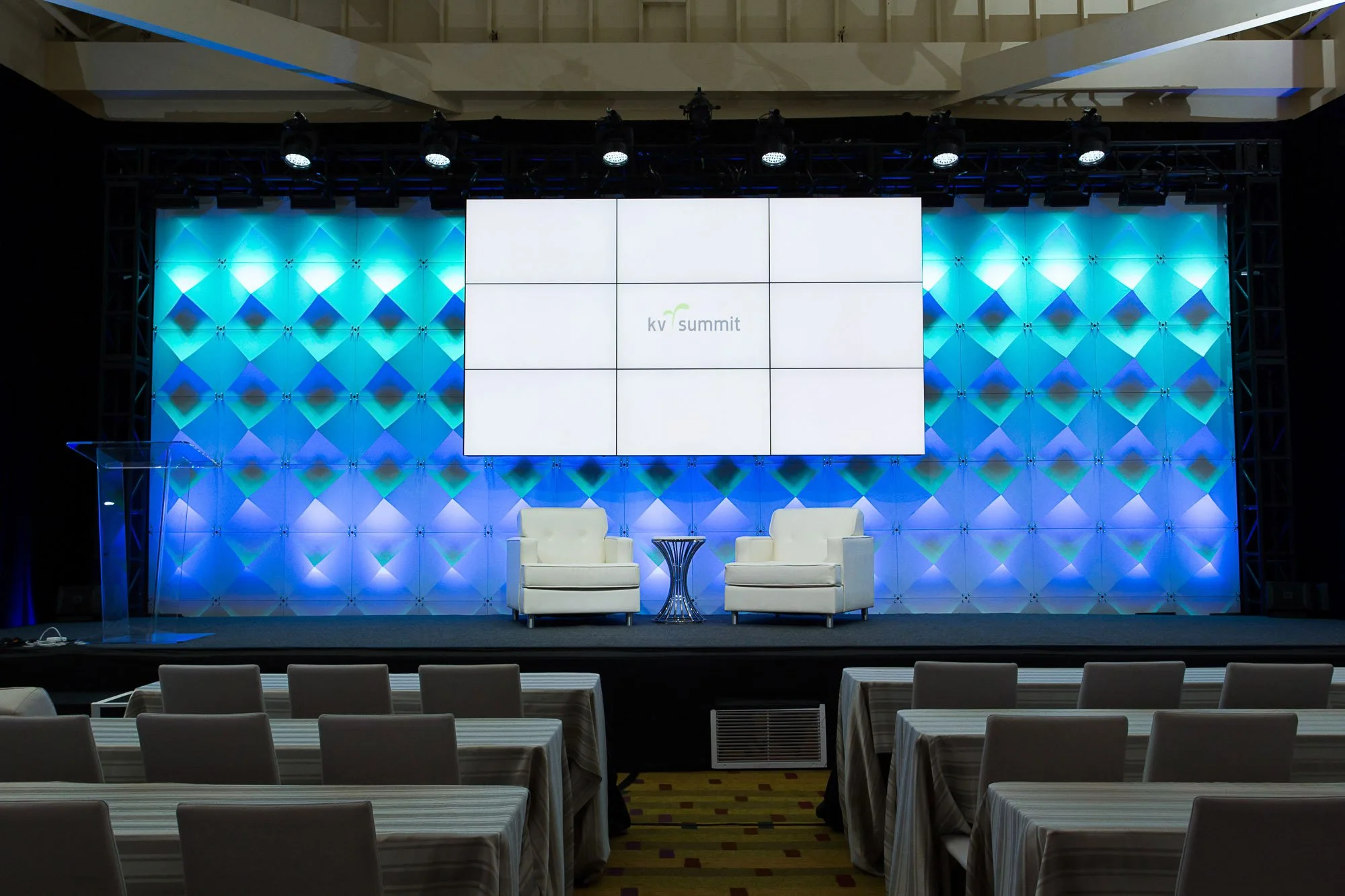 Stage with large screen displaying 'kv summit' logo, two white chairs and a small table, surrounded by blue lighting on a patterned wall in a conference room.