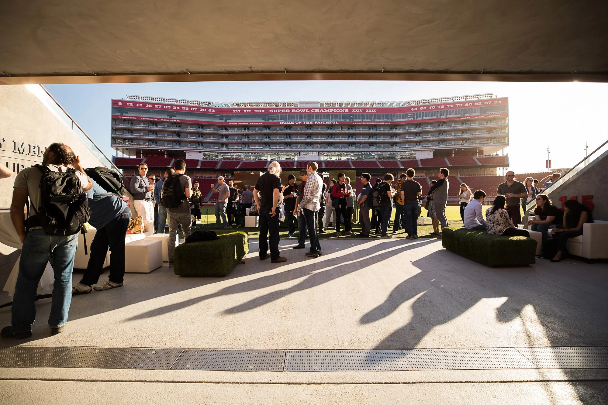 People gathered on a sports stadium field, some standing and talking, others sitting on benches, with stadium seating in the background, under a partially shaded area.