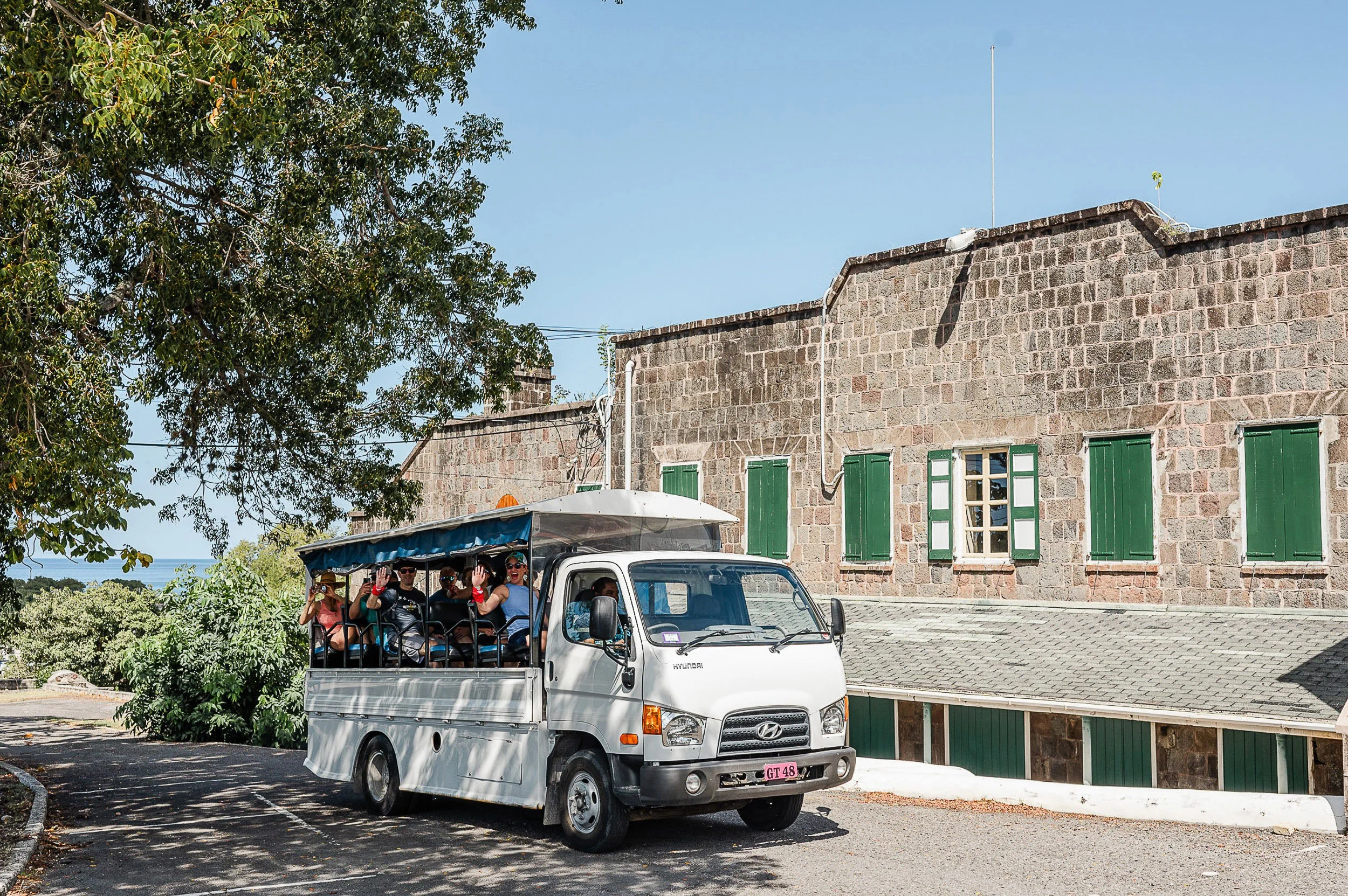 A white open-air bus with passengers smiling and waving, parked on a shaded street in front of a stone building with green window shutters. There is a large leafy tree on the left side and a clear blue sky above.