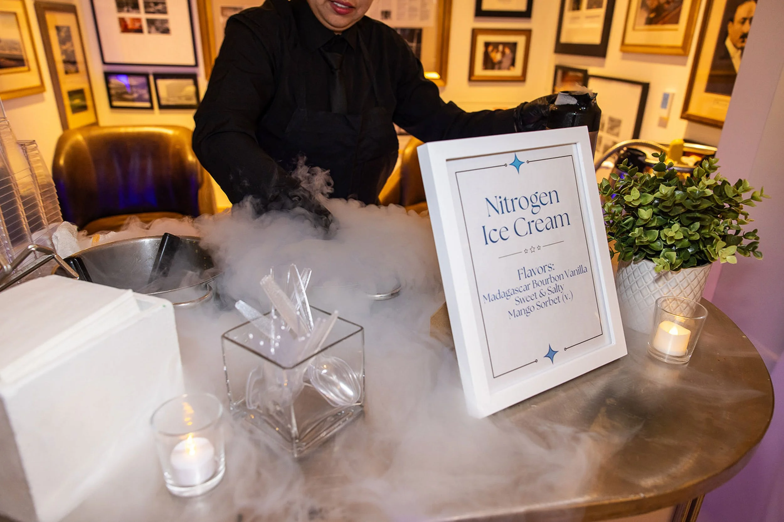 Person preparing nitrogen ice cream at a counter with a sign listing flavors. The person is wearing black gloves and a black apron, and there is dry ice smoke. The background has framed photos on the wall and a potted plant on the counter.