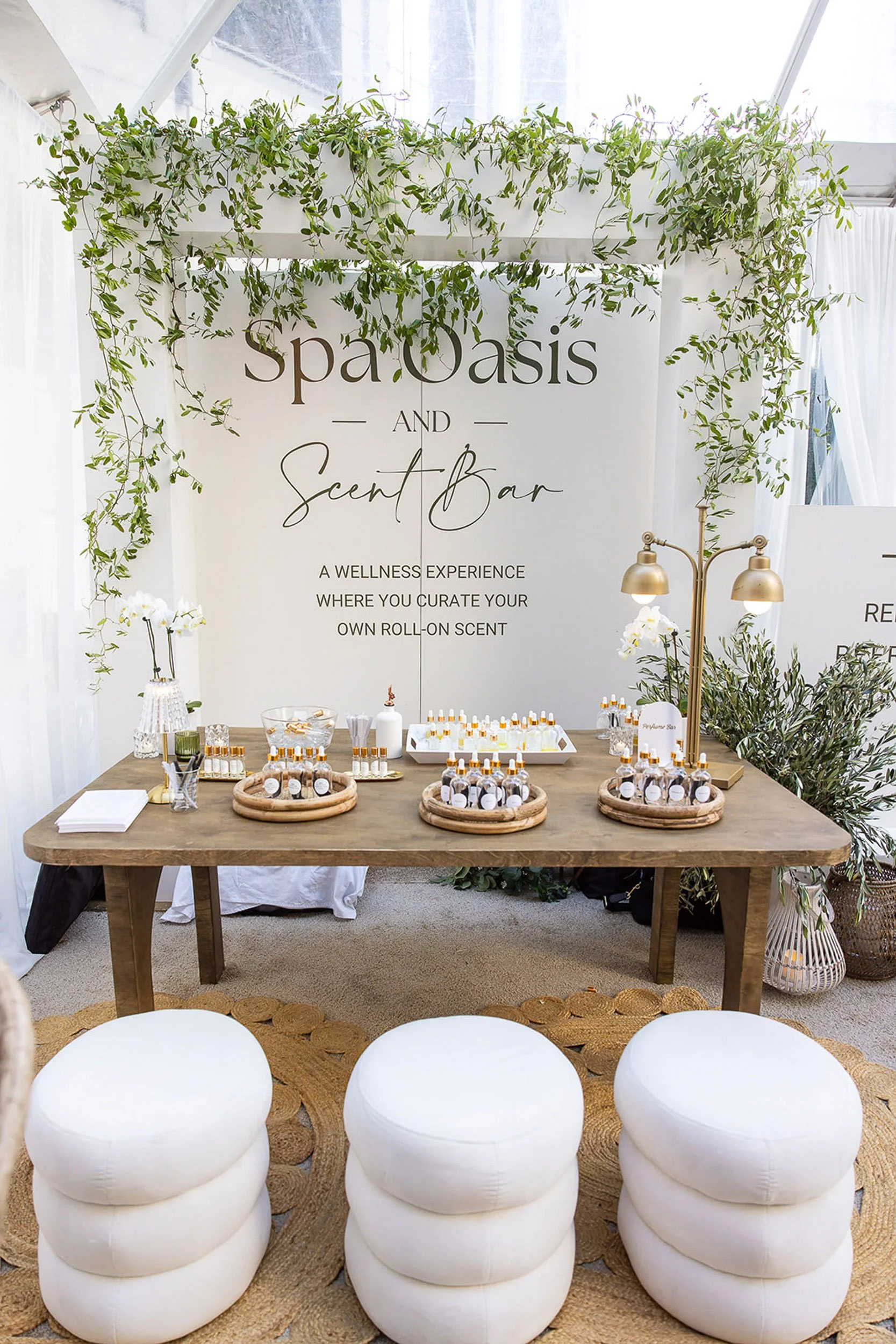 A table setup at an event with small bottles of scent or perfume, decorative elements, and a large sign that reads 'Spa Oasis and Scent Bar' with additional text about a wellness experience and custom scents. The area is decorated with greenery, whit