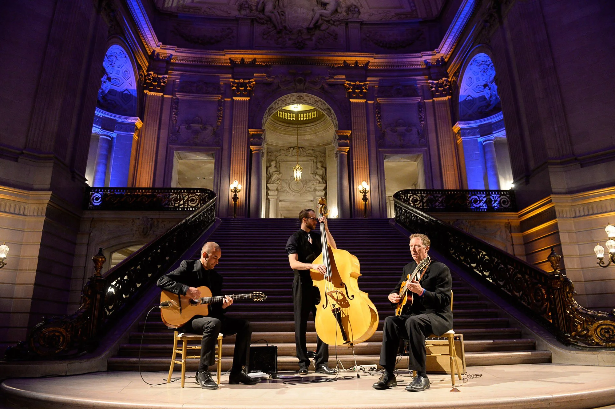 A jazz band performing on a grand staircase inside a historic building with ornate architecture and blue lighting. The band includes a guitarist, a double bassist, and a guitarist, all dressed in black.