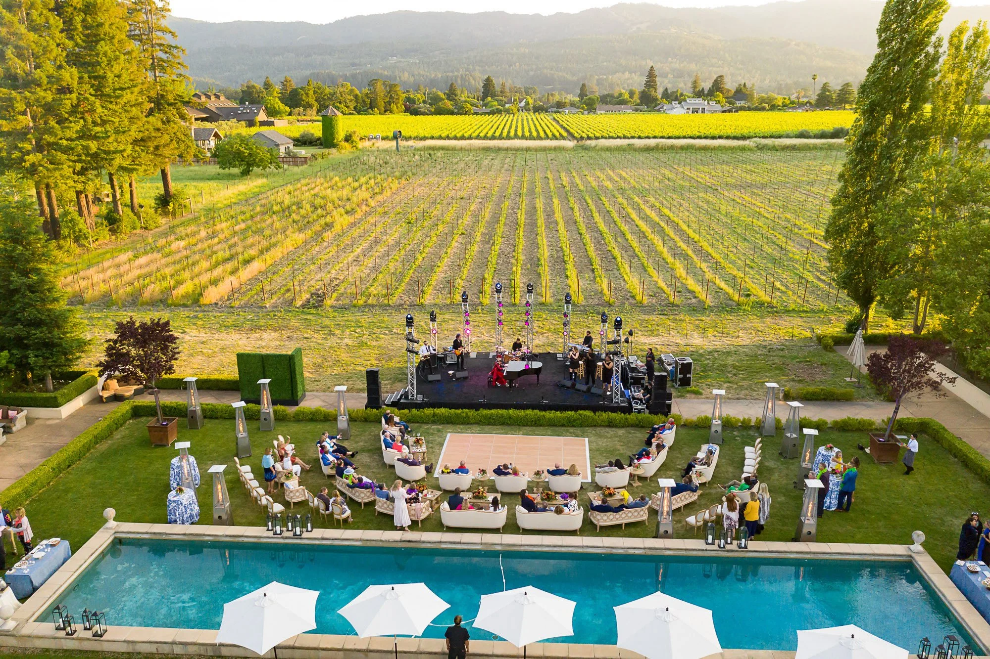 An outdoor concert is taking place in a backyard with a swimming pool. The audience is seated in white chairs and sofas, watching a band perform on a small stage. The setting is lush with trees, a vineyard in the background, and mountains beyond. The