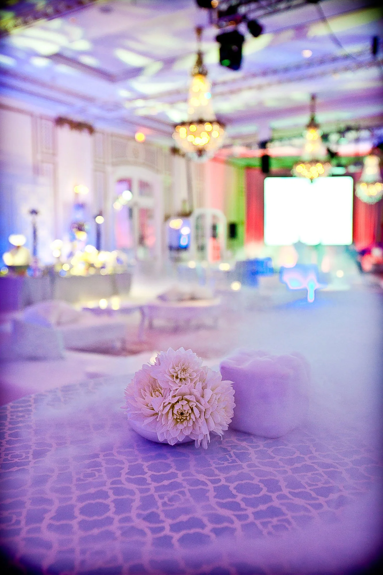 A blurred indoor event space with chandeliers, spotlights, and a large bright screen. In the foreground, a white table with a pink flower centerpiece and purple patterned tablecloth.