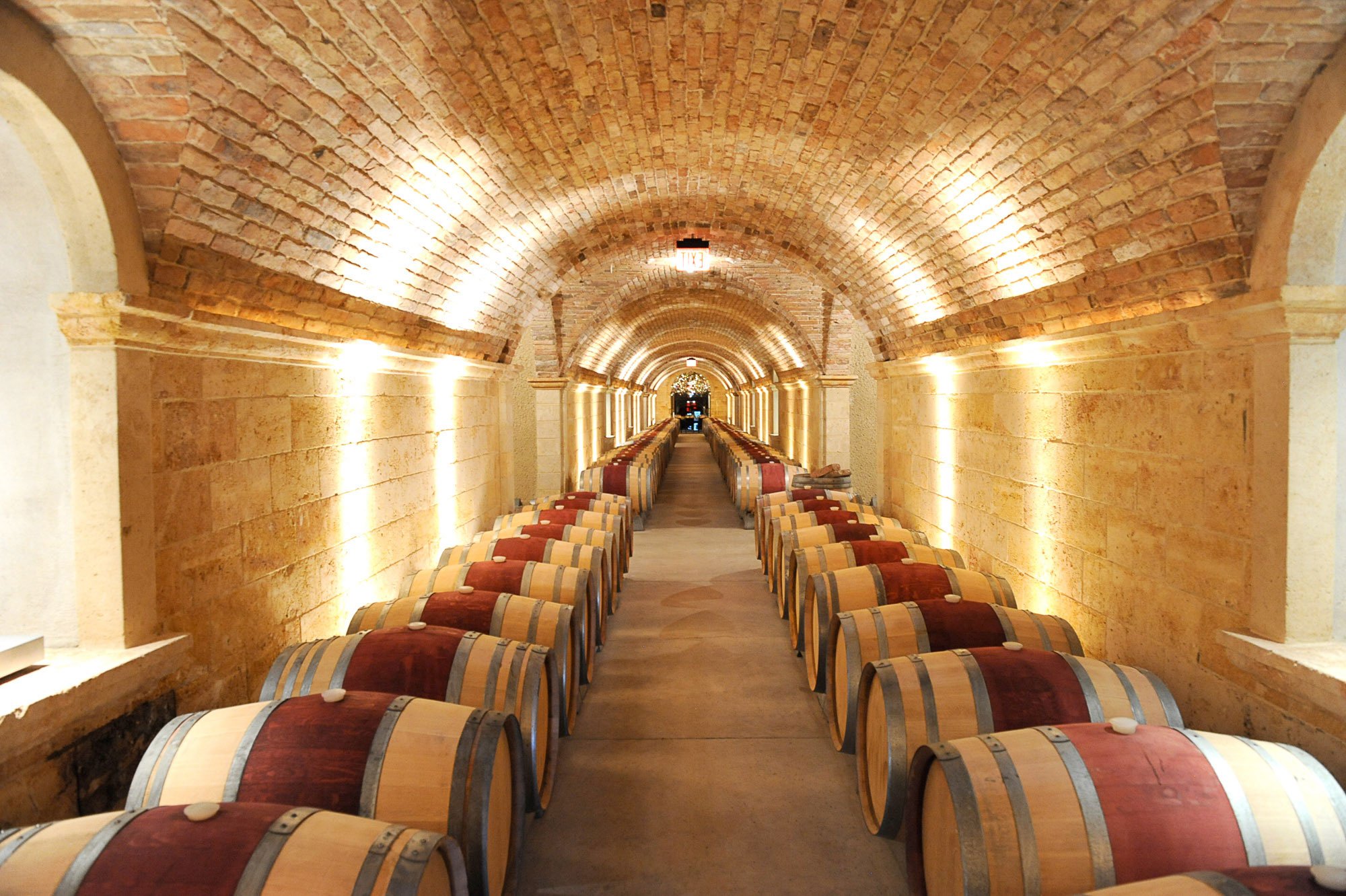 Underground wine cellar with arched brick ceiling, stone walls, arranged wine barrels, and soft lighting.