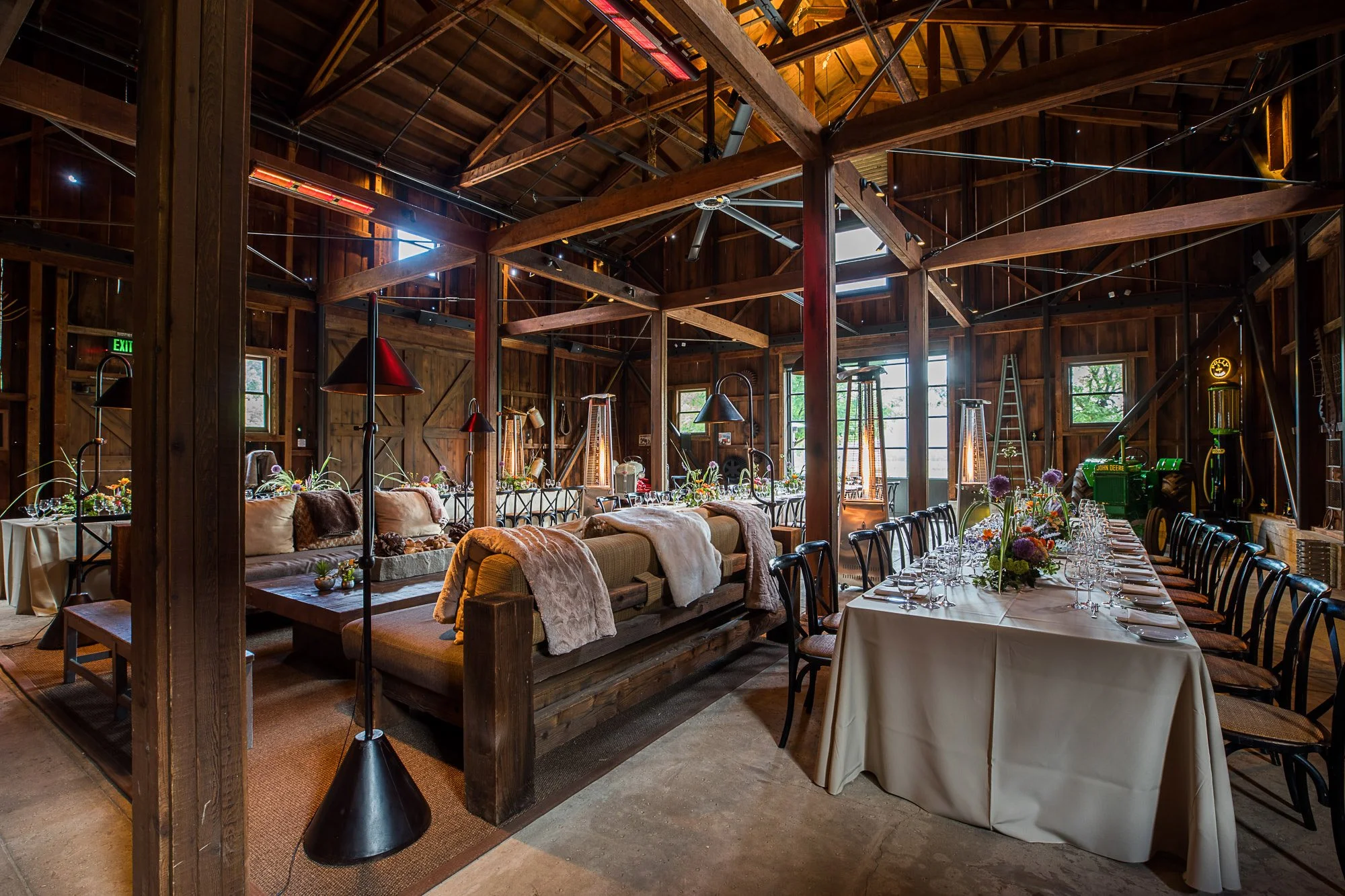 Interior of a rustic wooden barn converted into a dining or event space with a long table set with glassware and floral centerpieces, cozy sofas, and heaters, featuring exposed wooden beams and large windows letting in natural light.