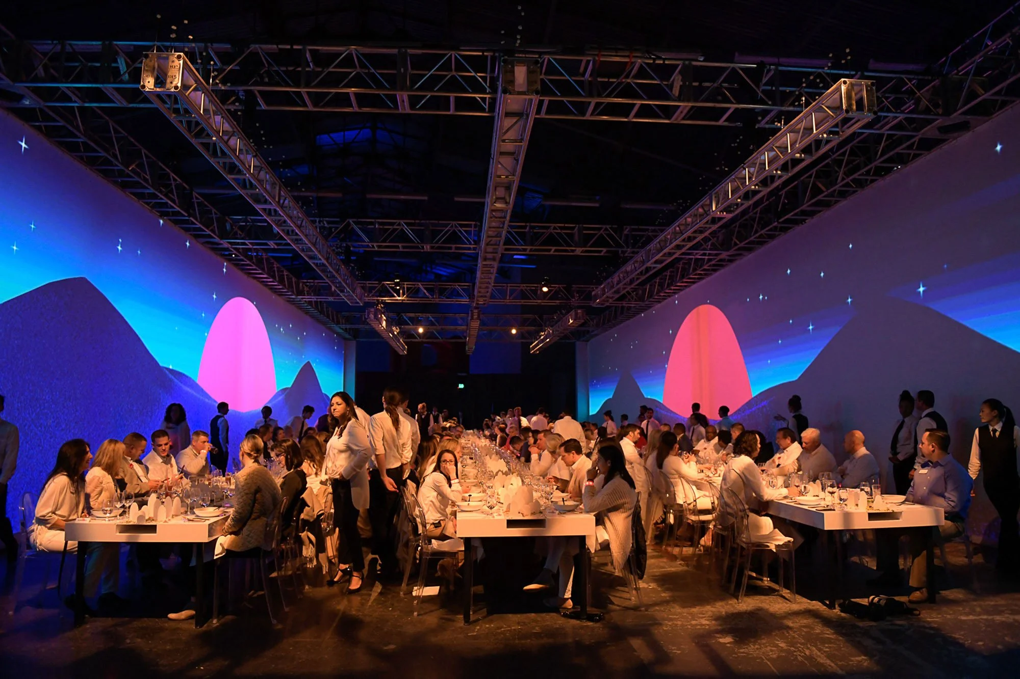 A large event space with high ceilings and elaborate lighting. Long banquet tables filled with seated people in white attire, possibly a formal dinner or conference. The background features a mountain landscape and starry sky projected on the walls.