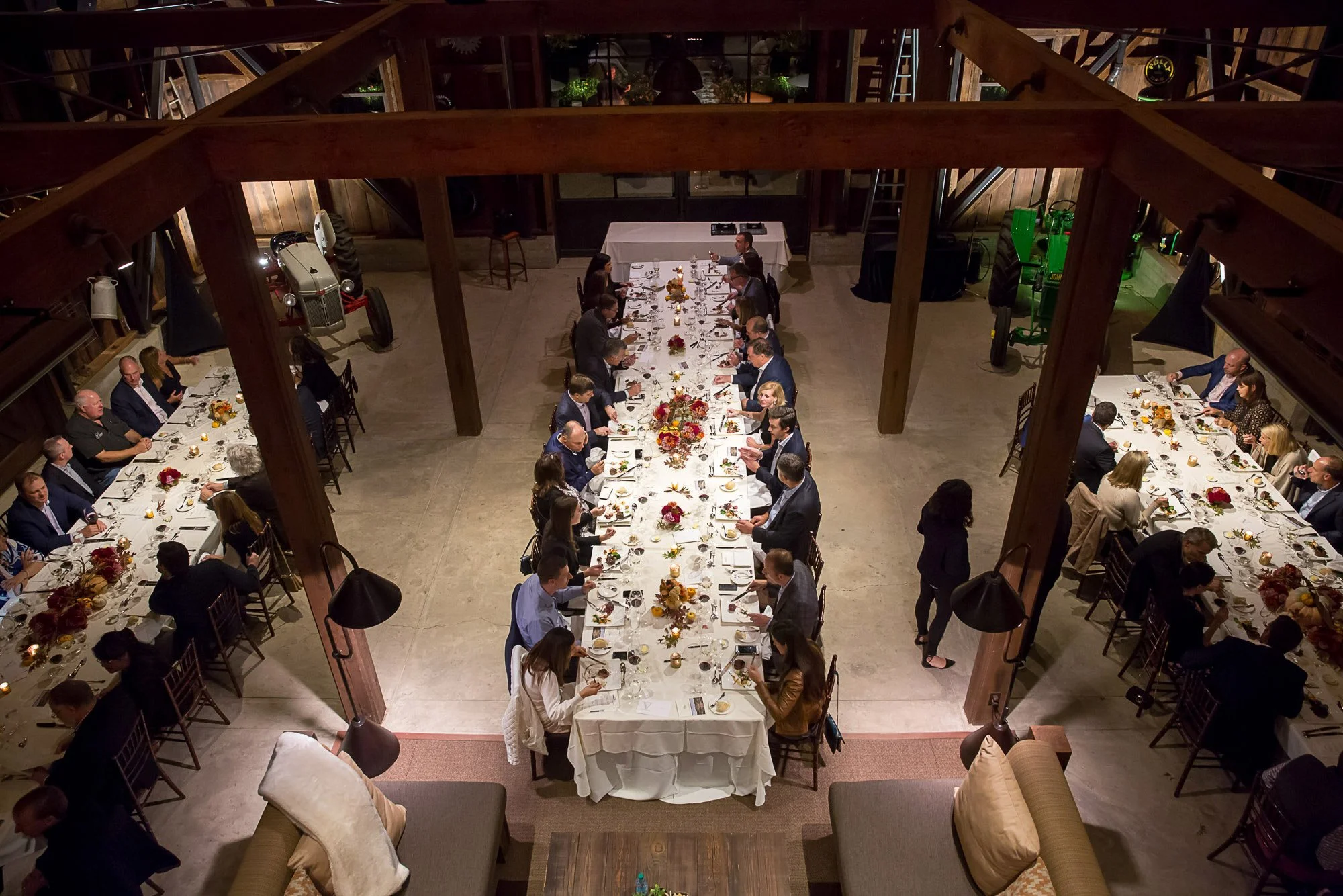A large group of people sitting at a long banquet table, with floral centerpieces and table settings, in a rustic venue with wooden beams and industrial decor.
