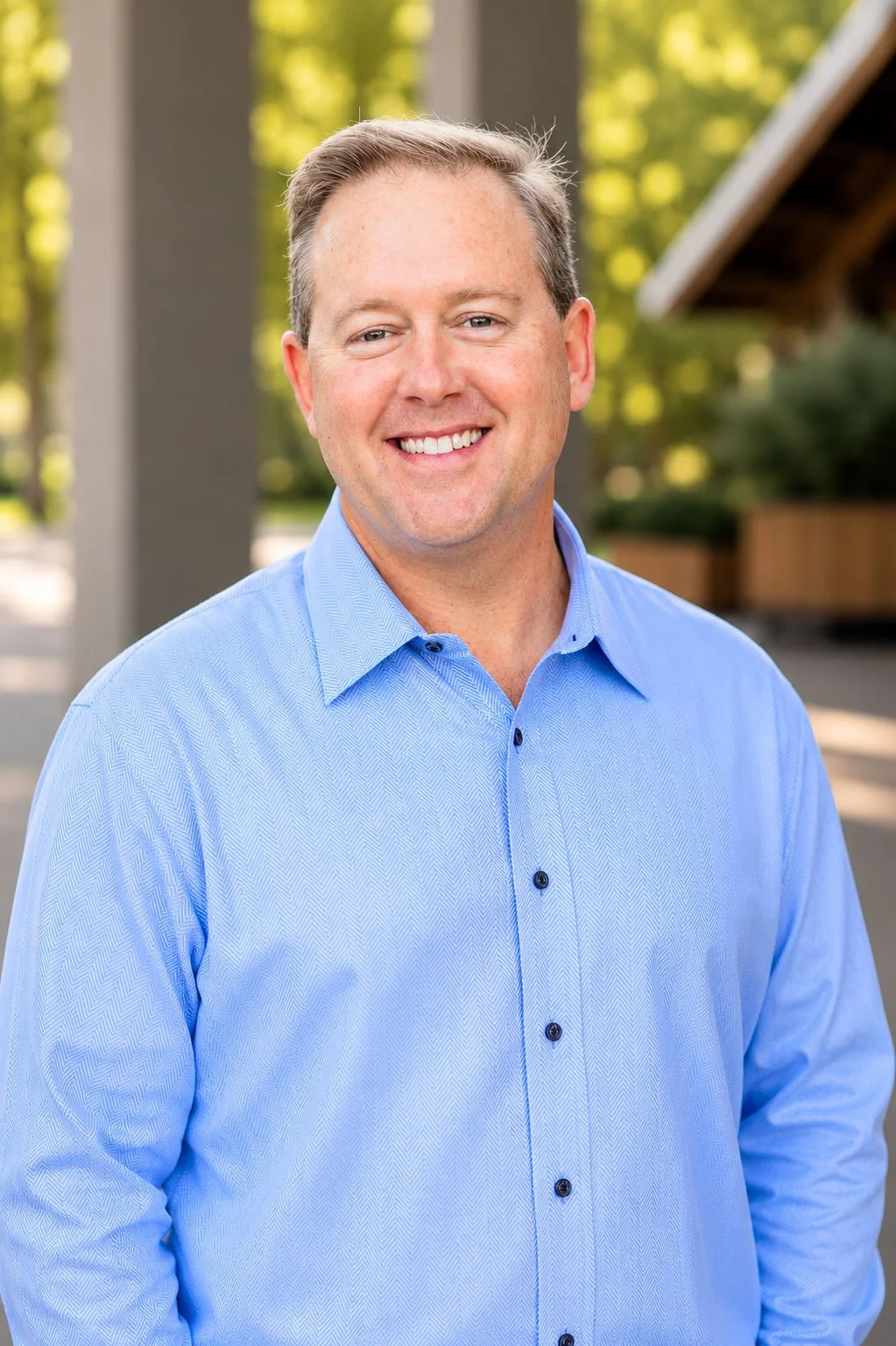 A smiling man with short light brown hair wearing a light blue button-up shirt standing outdoors with trees and a building in the background.