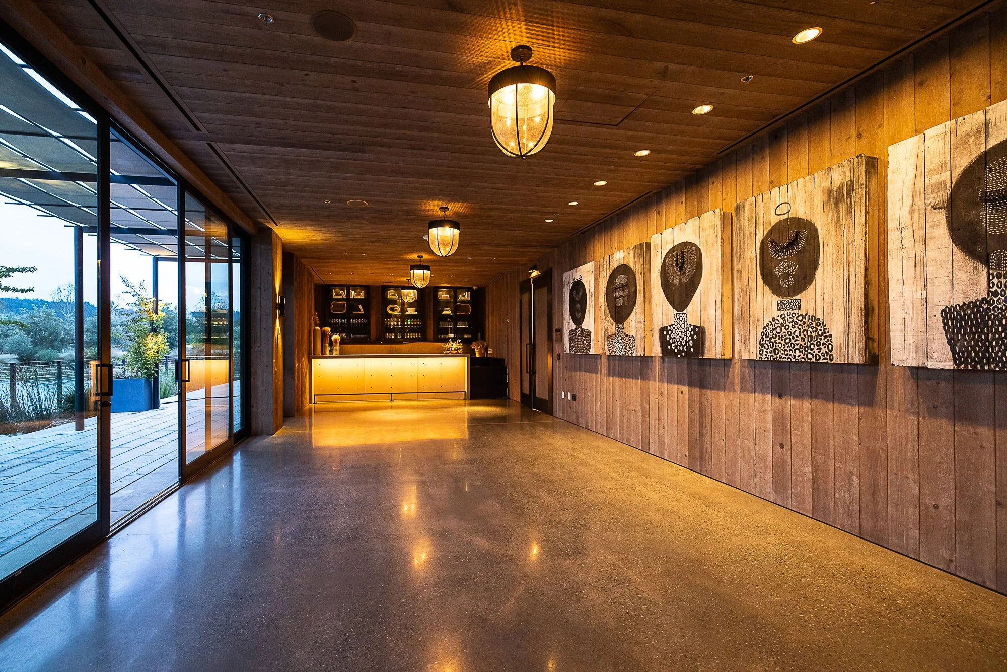 Interior of a contemporary building with wooden walls and ceiling, art on walls, large glass doors leading outside, modern light fixtures, and a polished concrete floor reflecting warm lighting.