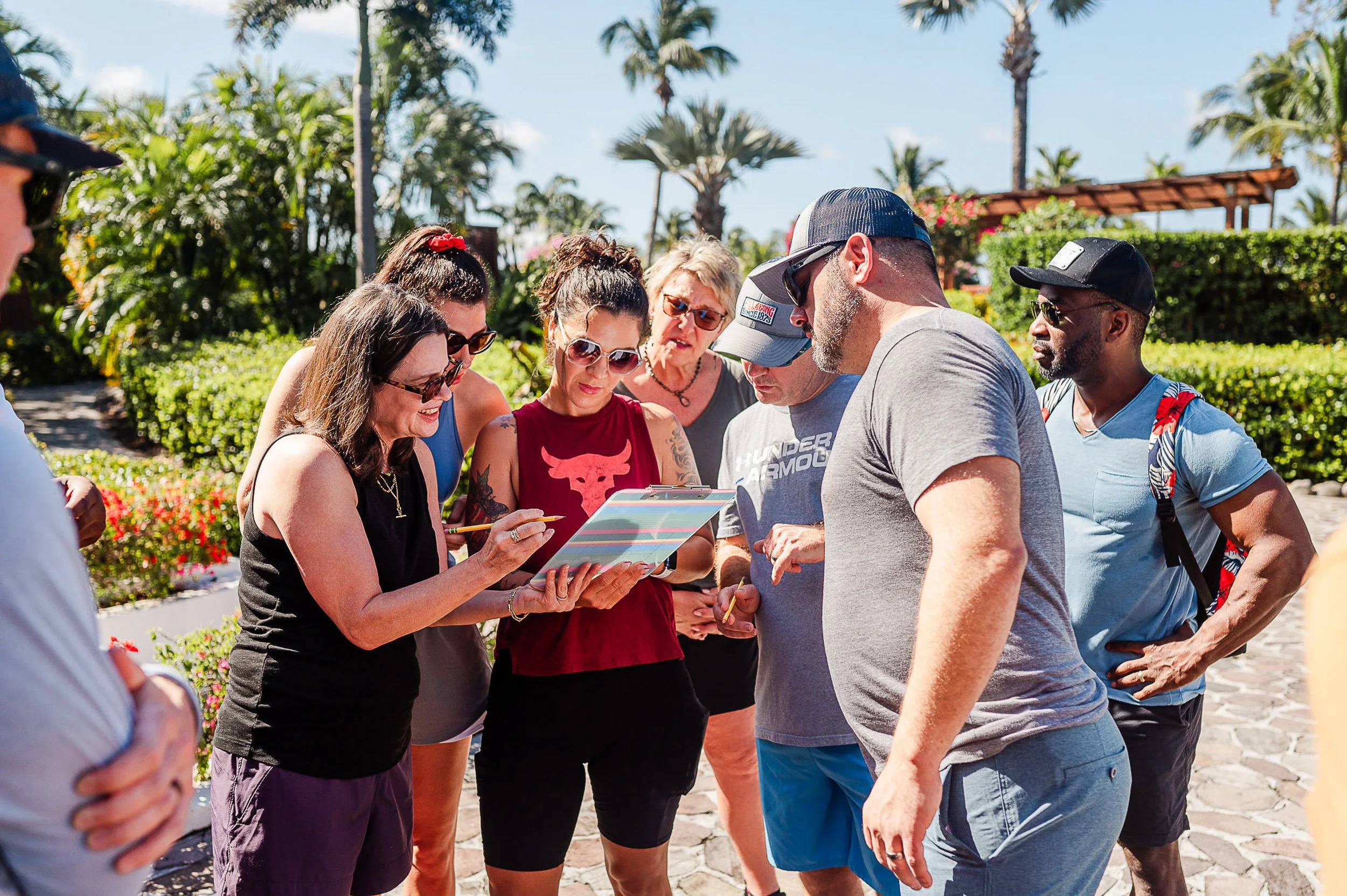 A group of adults gathered outdoors on a sunny day, looking at a clipboard or tablet held by a woman with sunglasses, surrounded by lush greenery and palm trees in the background.