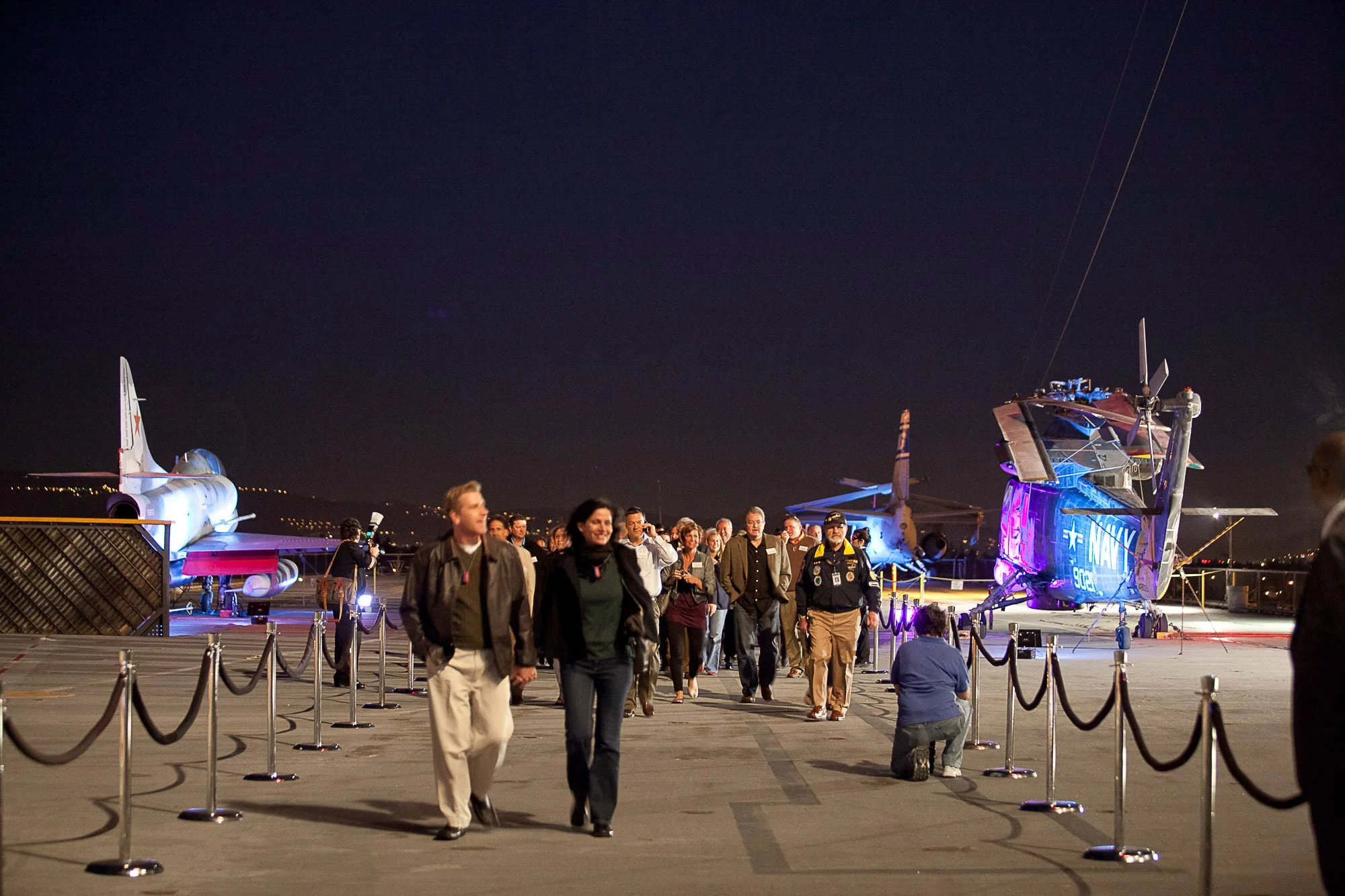 Nighttime scene on an aircraft carrier deck with a crowd of people walking along lined-up barriers. In the background are military aircraft, including a fighter jet and a helicopter, illuminated with colorful lights.