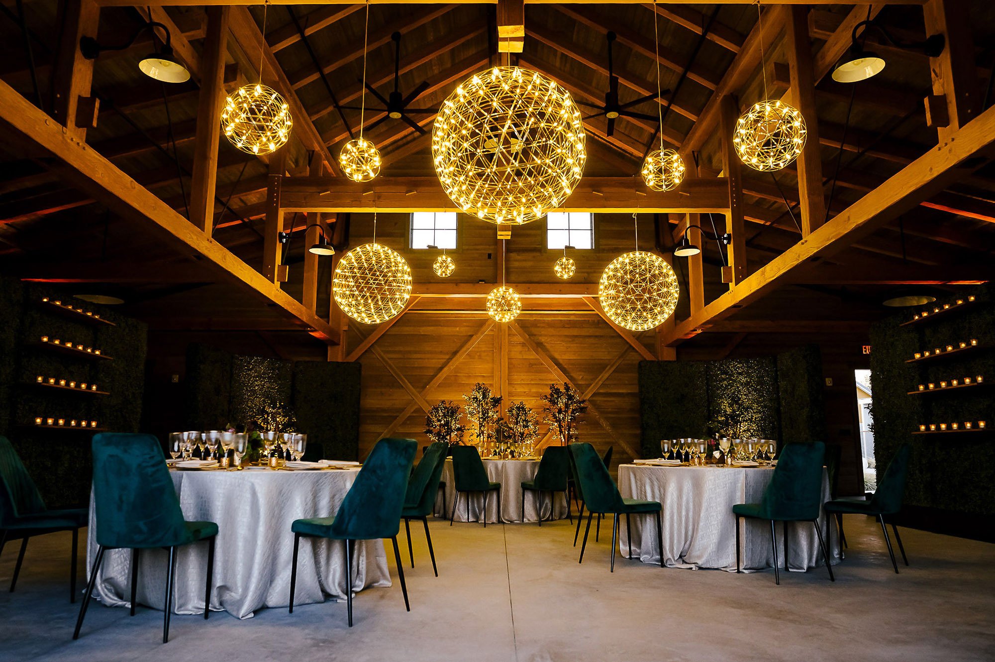 Interior of a rustic event hall with wooden beams, decorated with large spherical pendant lights, tables with white tablecloths, green chairs, and centerpieces, set for a formal event.