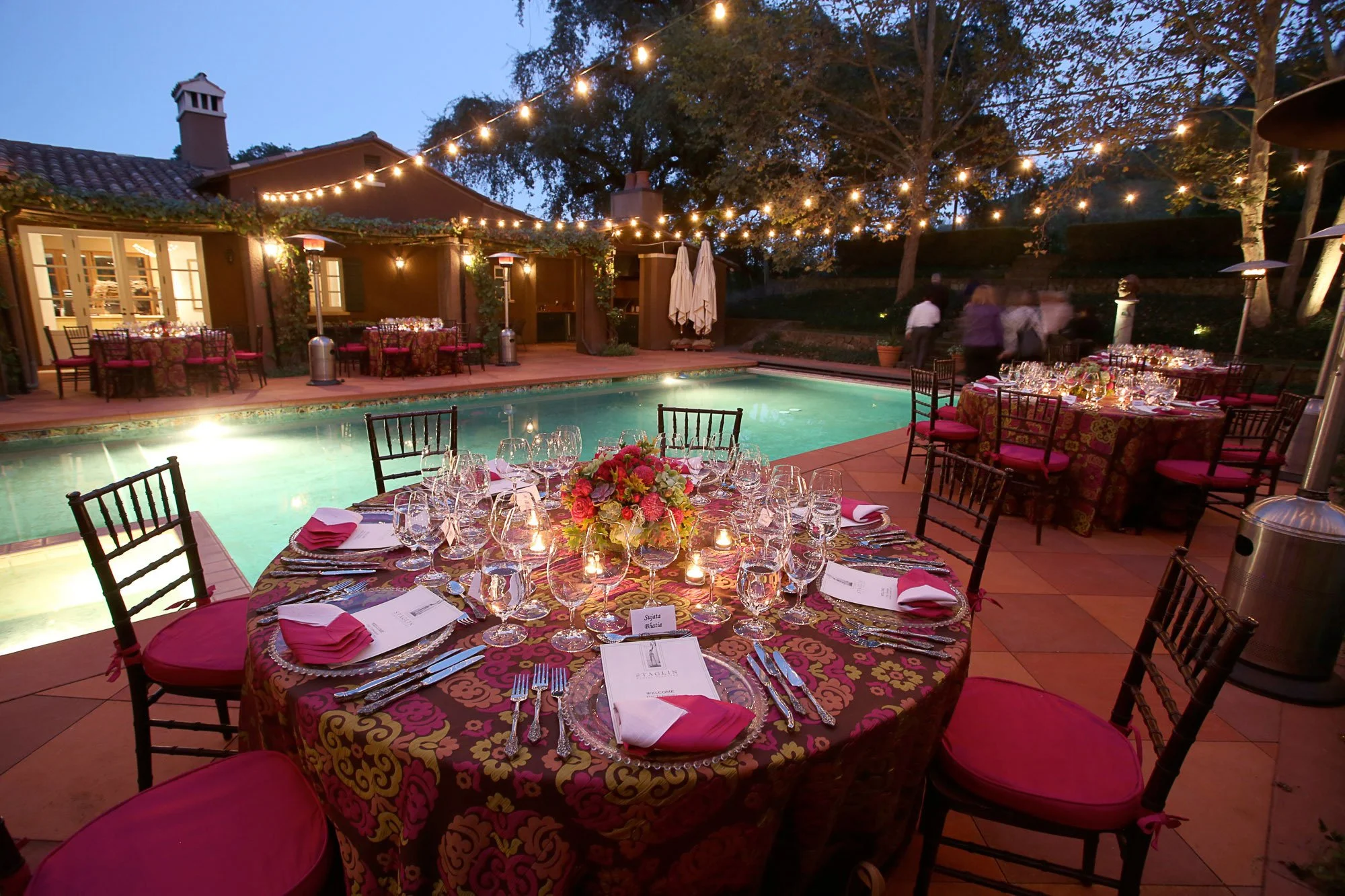 Elegant outdoor dinner setup with tables covered in patterned cloths, decorated with multiple wine glasses, candles, and a floral centerpiece, situated beside a swimming pool under string lights with a house and trees in the background during dusk.
