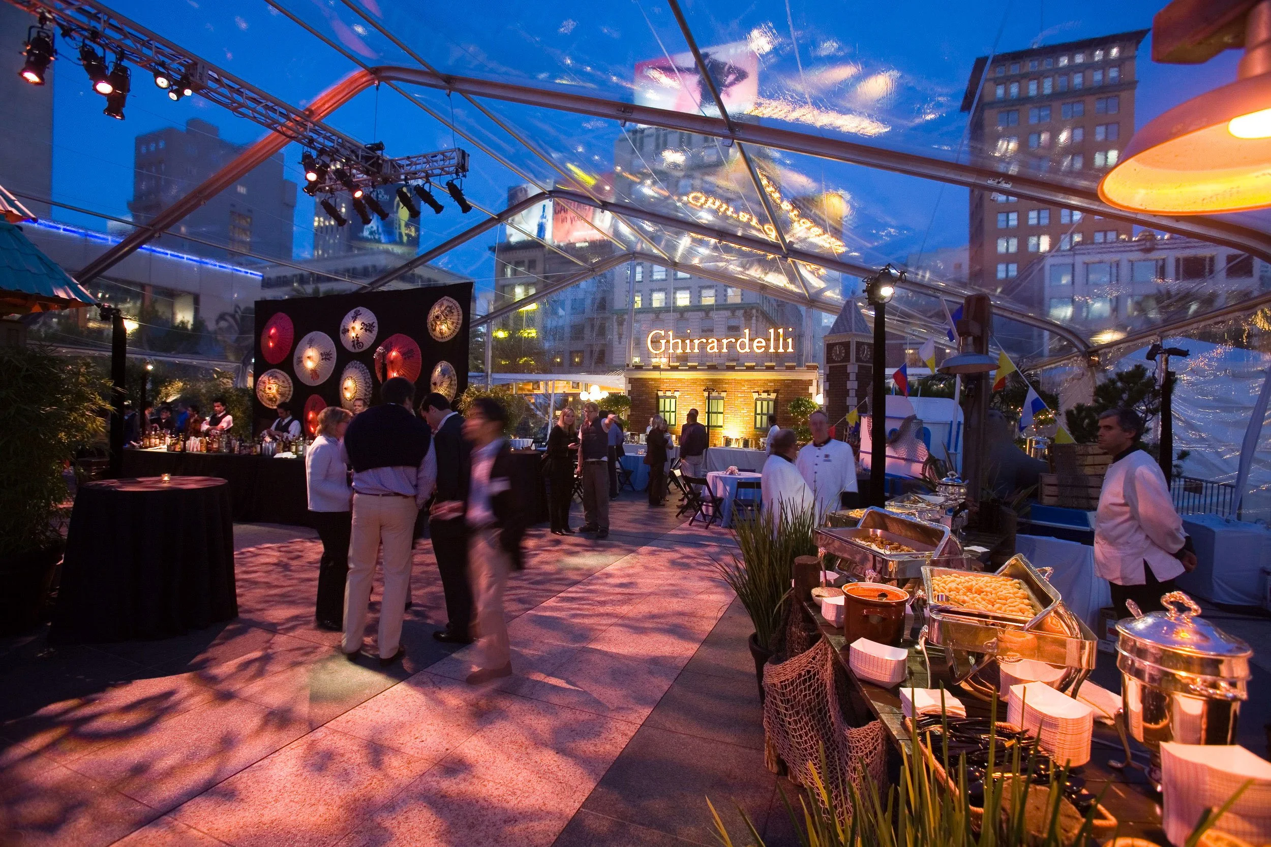 An outdoor evening event under a transparent tent with string lights and city buildings in the background. There are people socializing, a bar setup, and food stations with buffet-style offerings, including salads and appetizers. A sign that says 'Gh