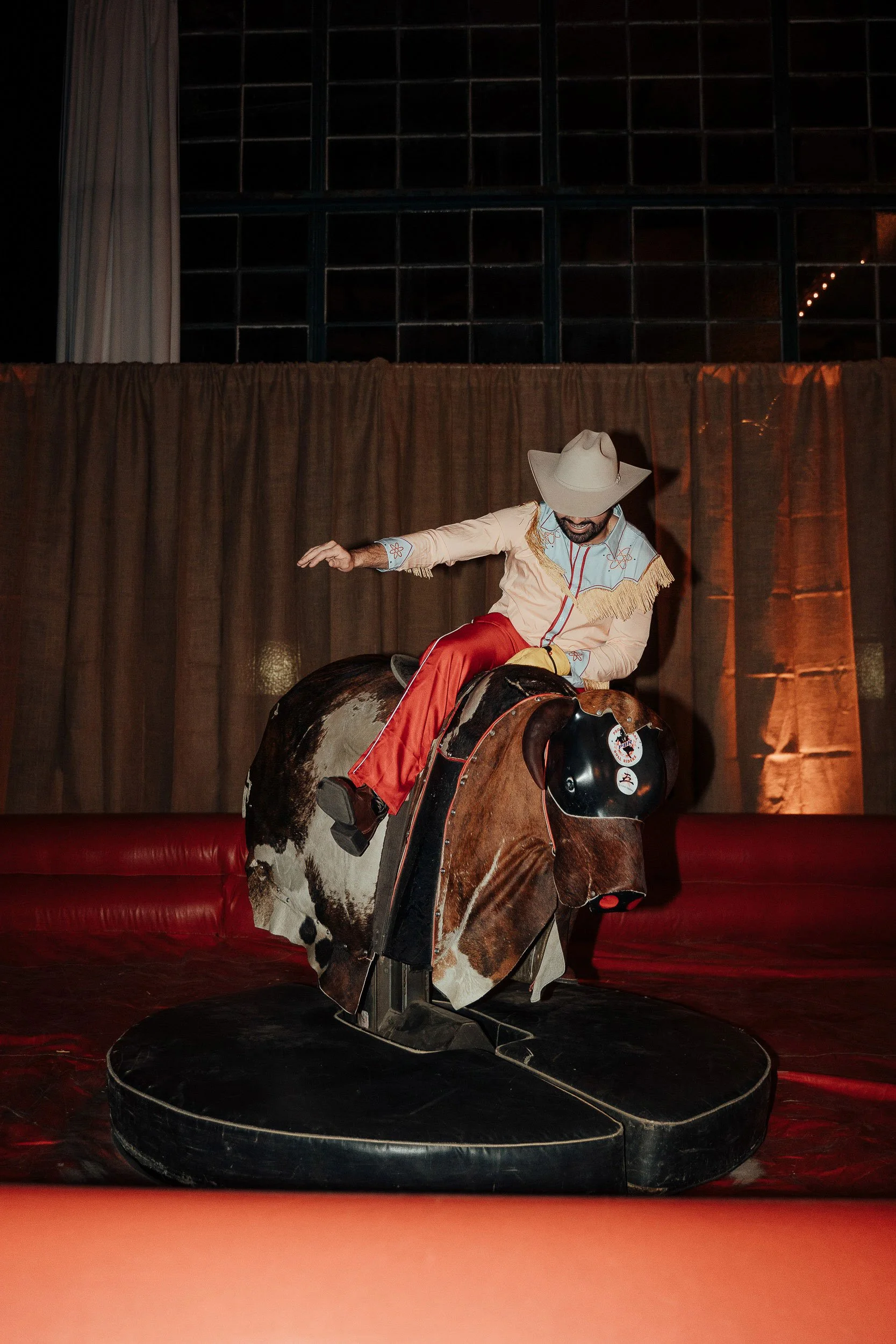 A man dressed as a cowboy, wearing a white hat and western attire, is riding a mechanical bull indoors. The background includes a brown curtain and large windows.