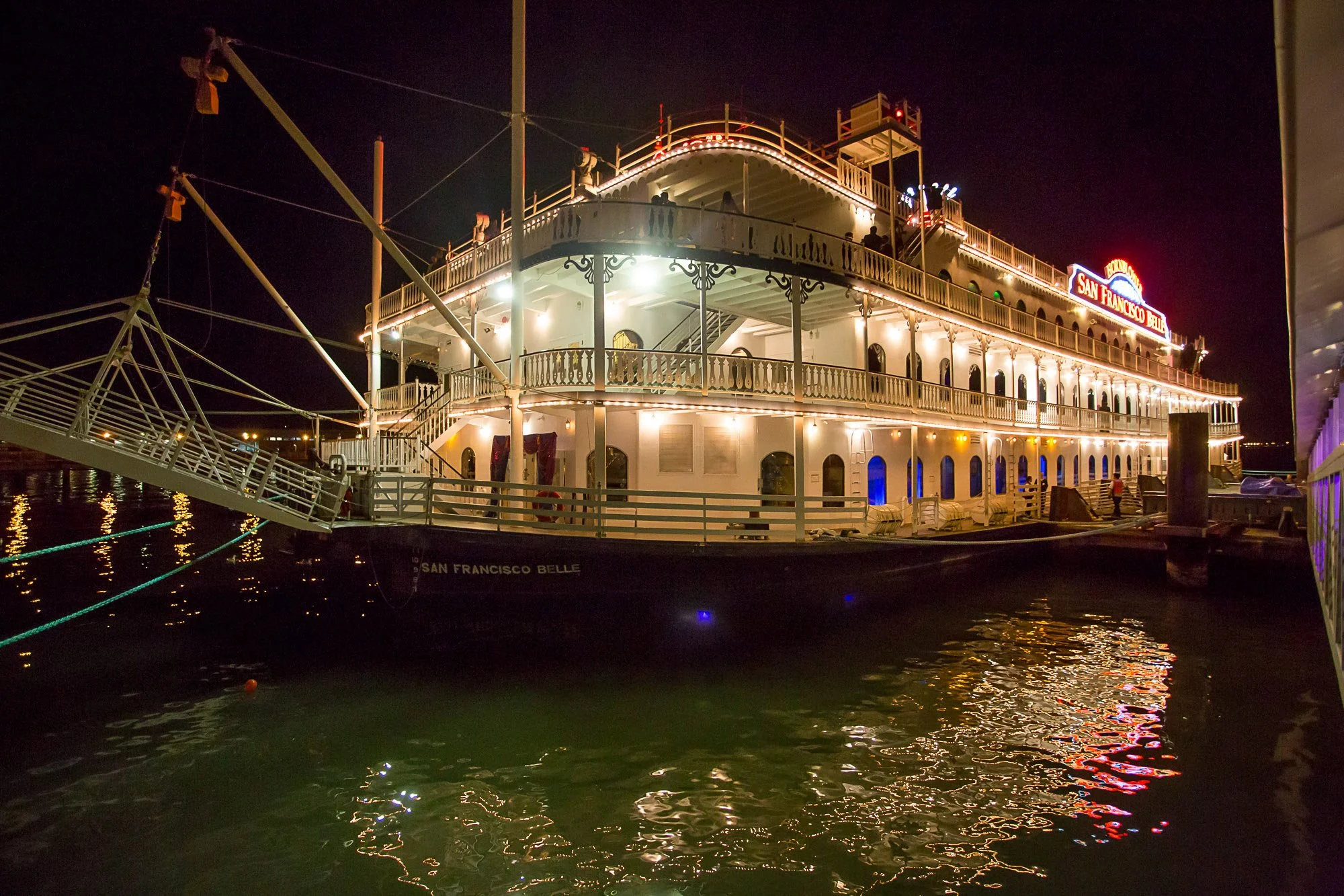 Night view of the San Francisco Belle paddleboat docked at the pier, illuminated with lights reflecting on the water, with a dark sky in the background.