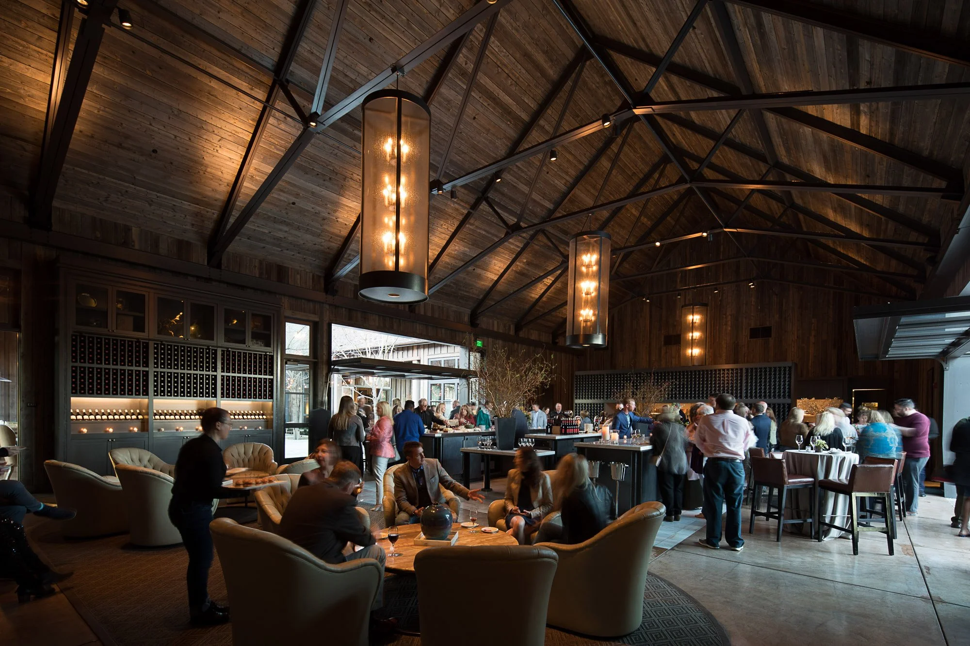 Indoor event space filled with people socializing, some seated and some standing, with tables, chairs, and wine glasses. The room has a high wooden ceiling with modern hanging light fixtures and large windows letting in natural light.