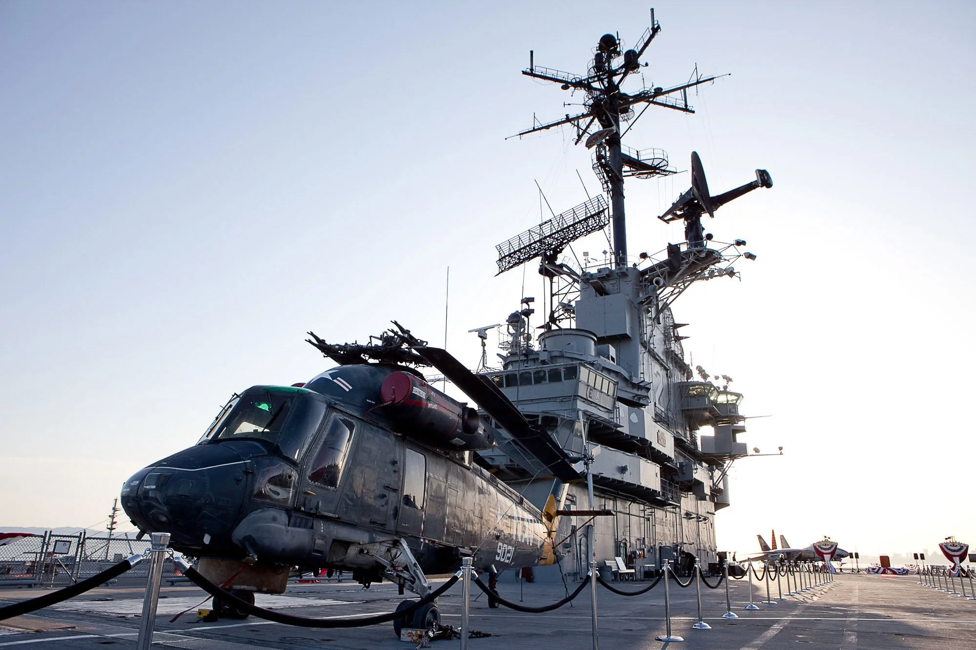 The photo shows a military aircraft carrier with a helicopter parked on the deck, surrounded by safety barriers, under a clear sky.