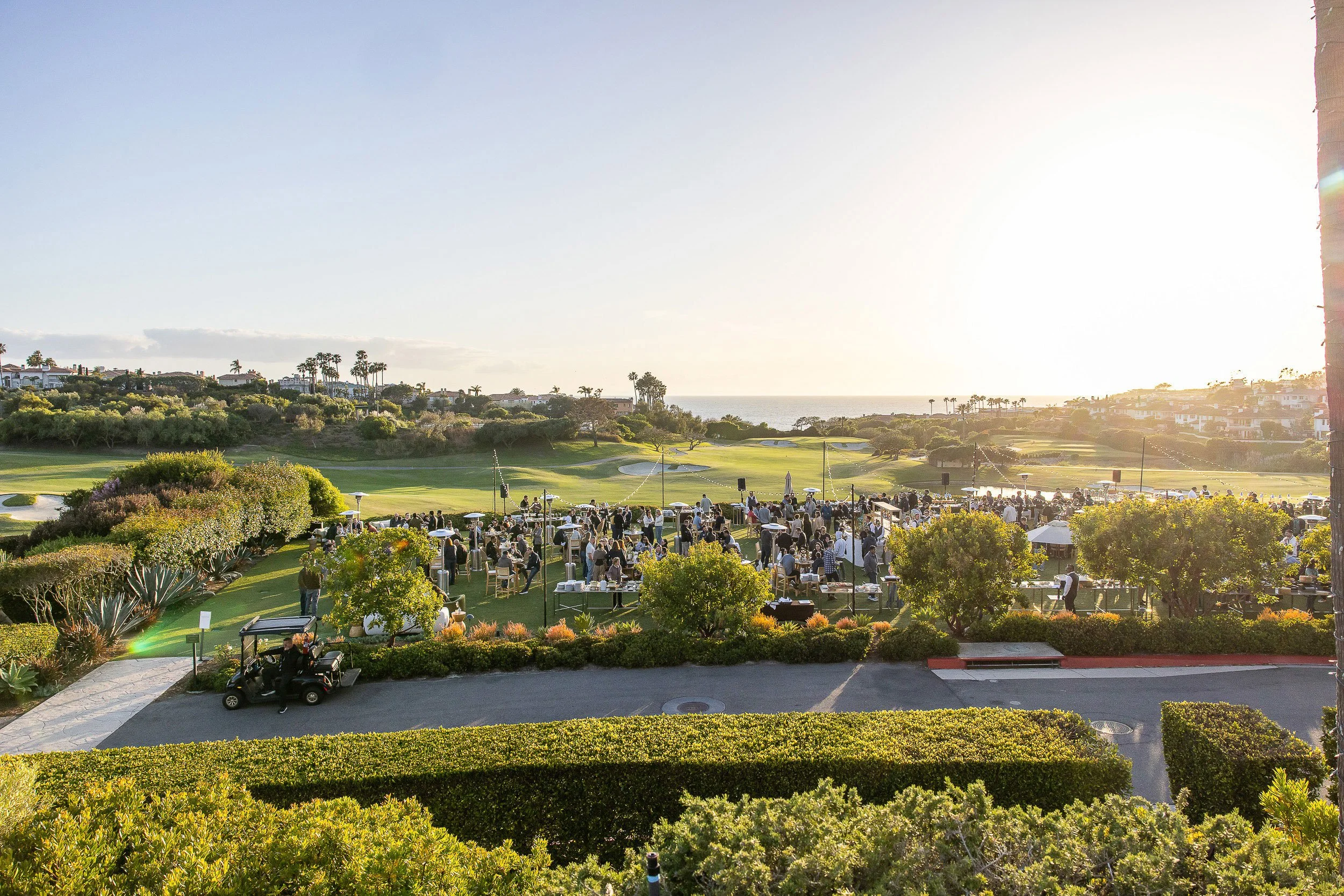 A scenic view of a golf course with lush green grass, trees, and a distant view of the ocean. There are many people gathered outdoors, some seated at tables and others standing, enjoying a social event in the late afternoon or early evening sunlight.