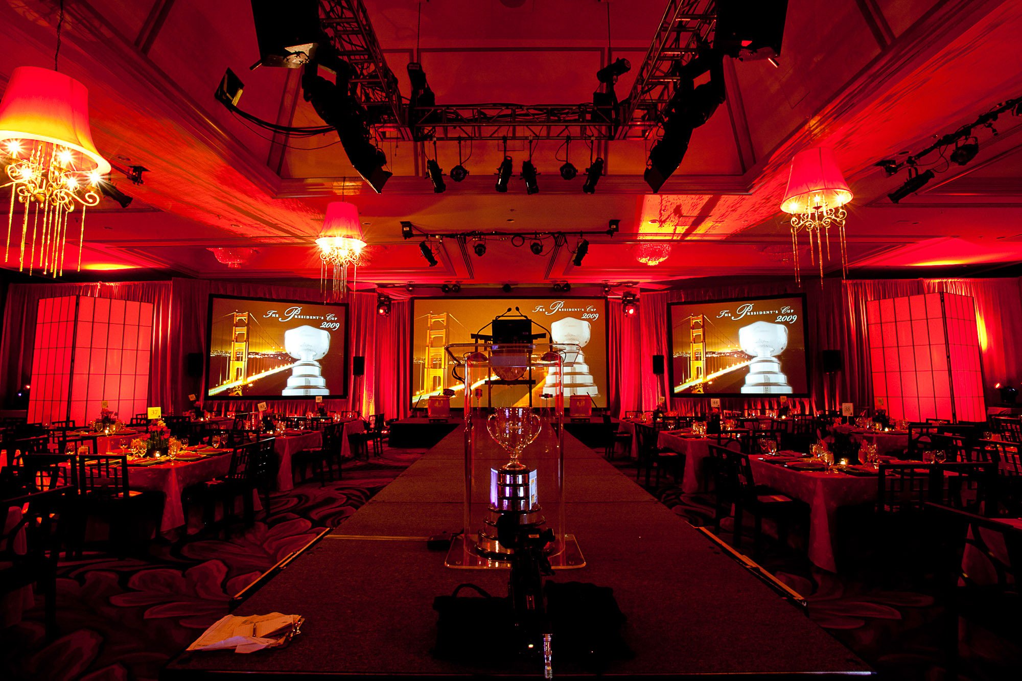 Banquet hall decorated for an event with red lighting, chandeliers, large screens displaying a trophy and the text "The President's Cup 2009," and empty tables with chairs and table settings.